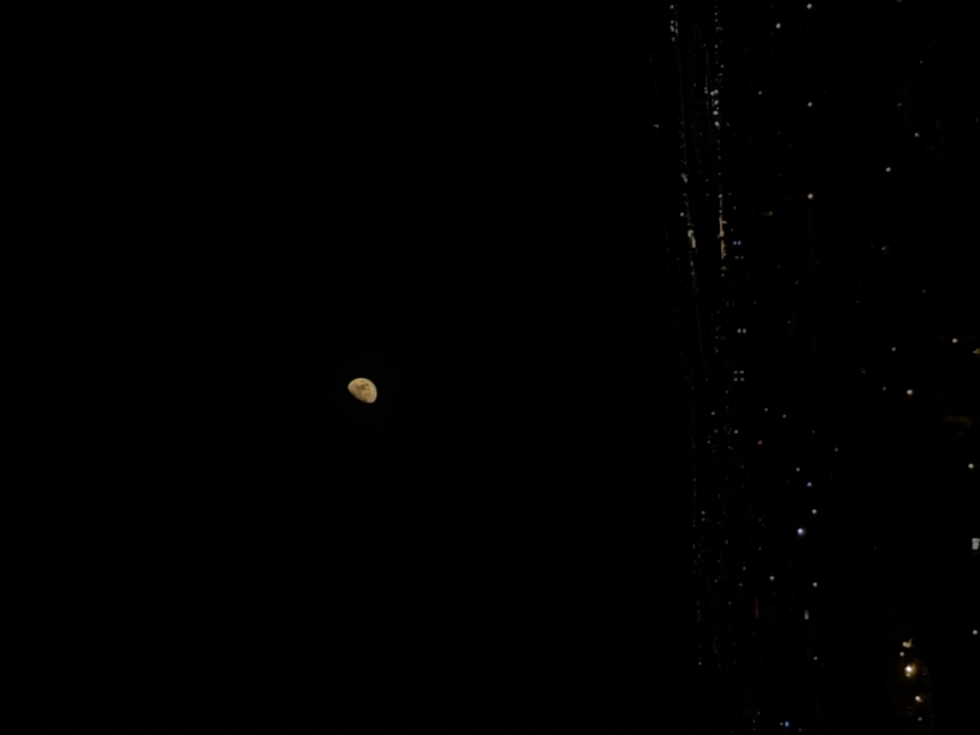A nearly full moon, tinged with yellow and showing some surface detail, is visible in the upper portion of a nearly black sky. Below the moon, a distant city skyline is illuminated by numerous small, scattered lights, suggesting a view over Konya, Turkey at night. The lights are predominantly white, with occasional hints of yellow and blue. The overall impression is one of a vast, dark urban landscape viewed from a distance under a nocturnal sky.