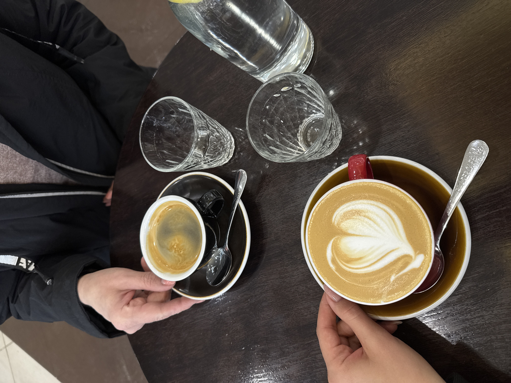 A high-angle shot shows a dark wooden table in an unidentified location in Unknown, Unknown. Two individuals are present, each holding a cup of coffee. The person on the left holds a small, white cup of espresso on a black saucer, with a silver spoon resting on the saucer. The person on the right holds a larger cup of latte with a heart-shaped foam design, in a tan-colored cup and saucer, also with a silver spoon. Between the two cups are two empty, crystal-cut glasses and a clear bottle of water. The person holding the espresso wears a dark jacket, while the other person wears a lighter garment.