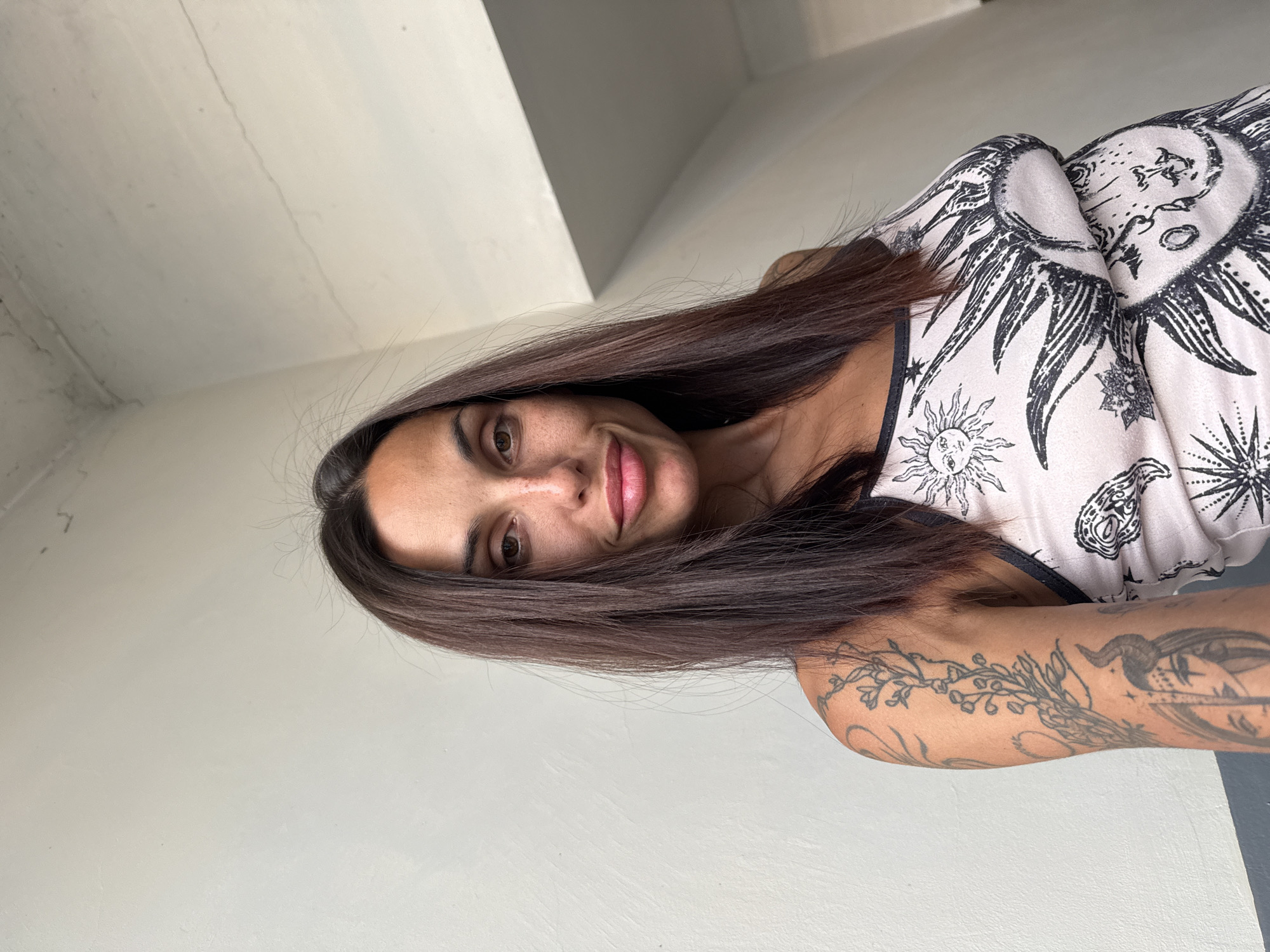 A woman with long, dark brown hair and freckles is looking at the camera. She is wearing a light-colored tank top with a sun-and-star pattern. She has tattoos on her left arm, including a floral design and a depiction of a face with horns. The background is a textured white wall with a concrete ceiling above. The image appears to have been taken indoors, possibly in a modern building. There is no discernible information in the image that would indicate a specific location within the city of Pleven, Bulgaria.