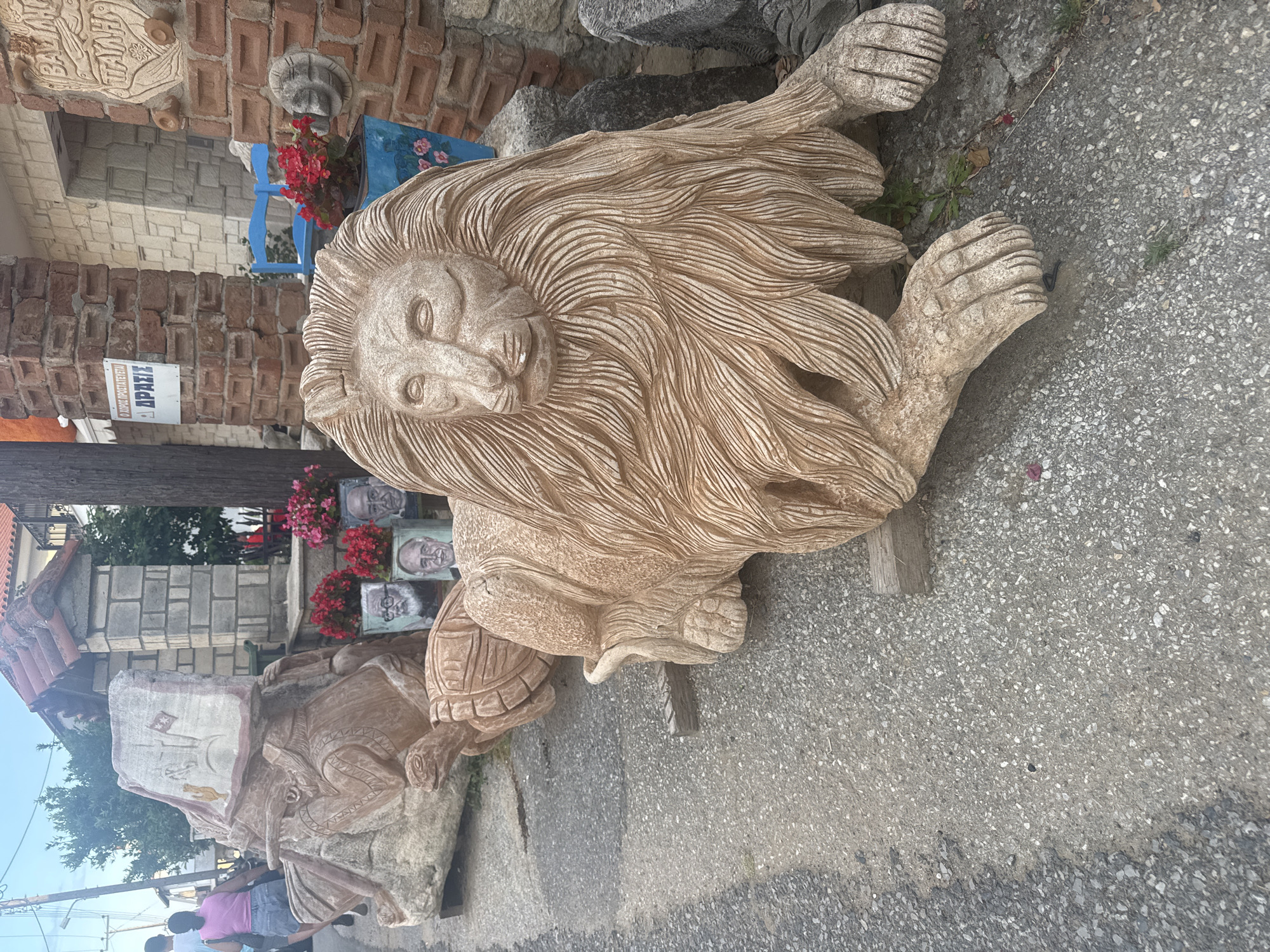 This image shows a street scene in the Kassandra Municipal Unit, Greece, featuring several stone sculptures. In the foreground, a large, light-brown stone sculpture of a lion is prominently displayed, lying down with its paws extended. Behind the lion, a turtle sculpture is partially visible. Further back, another large stone sculpture depicts a figure wearing a hat and holding a flag, with a smaller turtle sculpture beneath it.
The sculptures are placed on a paved surface, possibly a sidewalk or road, with some vegetation growing in cracks. In the background, there is a brick building with stone accents. A blue chair and potted plants with red flowers are visible near the building. A sign with Greek text is attached to a brick pillar. Two individuals, a woman in a pink top and denim shorts and another person, are walking away from the sculptures on the left side of the image. The lighting suggests it is daytime, likely late afternoon or early evening, given the soft light.