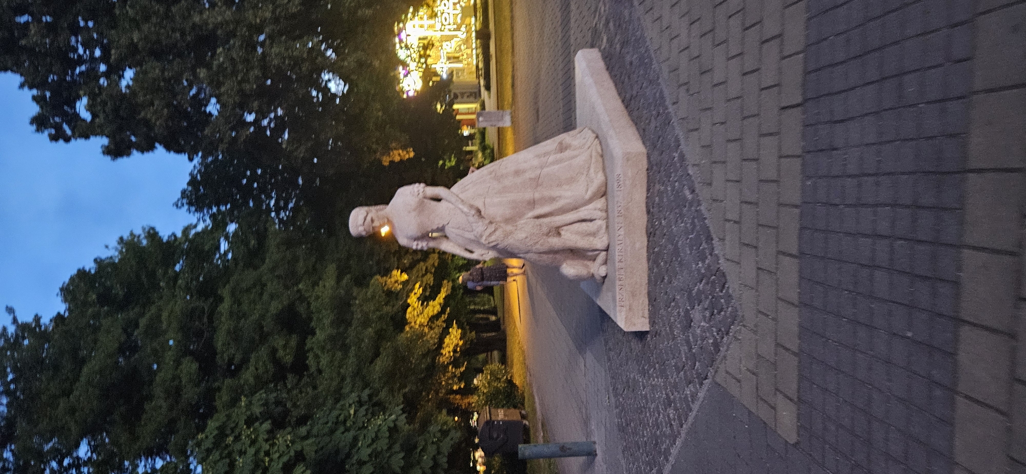 The image shows a statue of a woman with a large, flowing dress and a dog at her feet, located in Keszthely, Hungary. The statue is made of stone and is mounted on a stone base inscribed with "ERZSEBET KIRALYNÉ 1837 1898". The statue is situated in a park-like setting with trees and a paved walkway. In the background, there are streetlights and a few people walking. The sky is dark blue, indicating it is either dusk or early evening. The overall atmosphere is calm and peaceful.