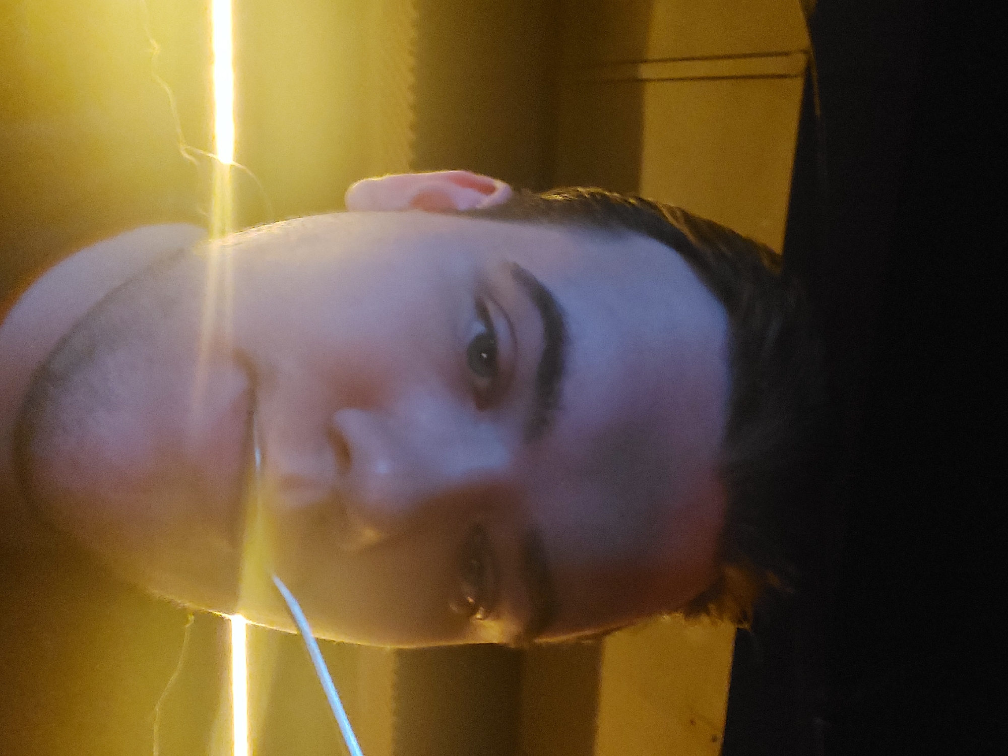 A close-up, rotated portrait of a person with fair skin. The subject has dark hair and visible stubble on the face. The background consists of yellow-toned lighting with vertical light streaks and dark, indistinct areas. The scene lacks specific details to indicate a location in Tashkent, Uzbekistan. There are no recognizable landmarks or cultural indicators within the frame.