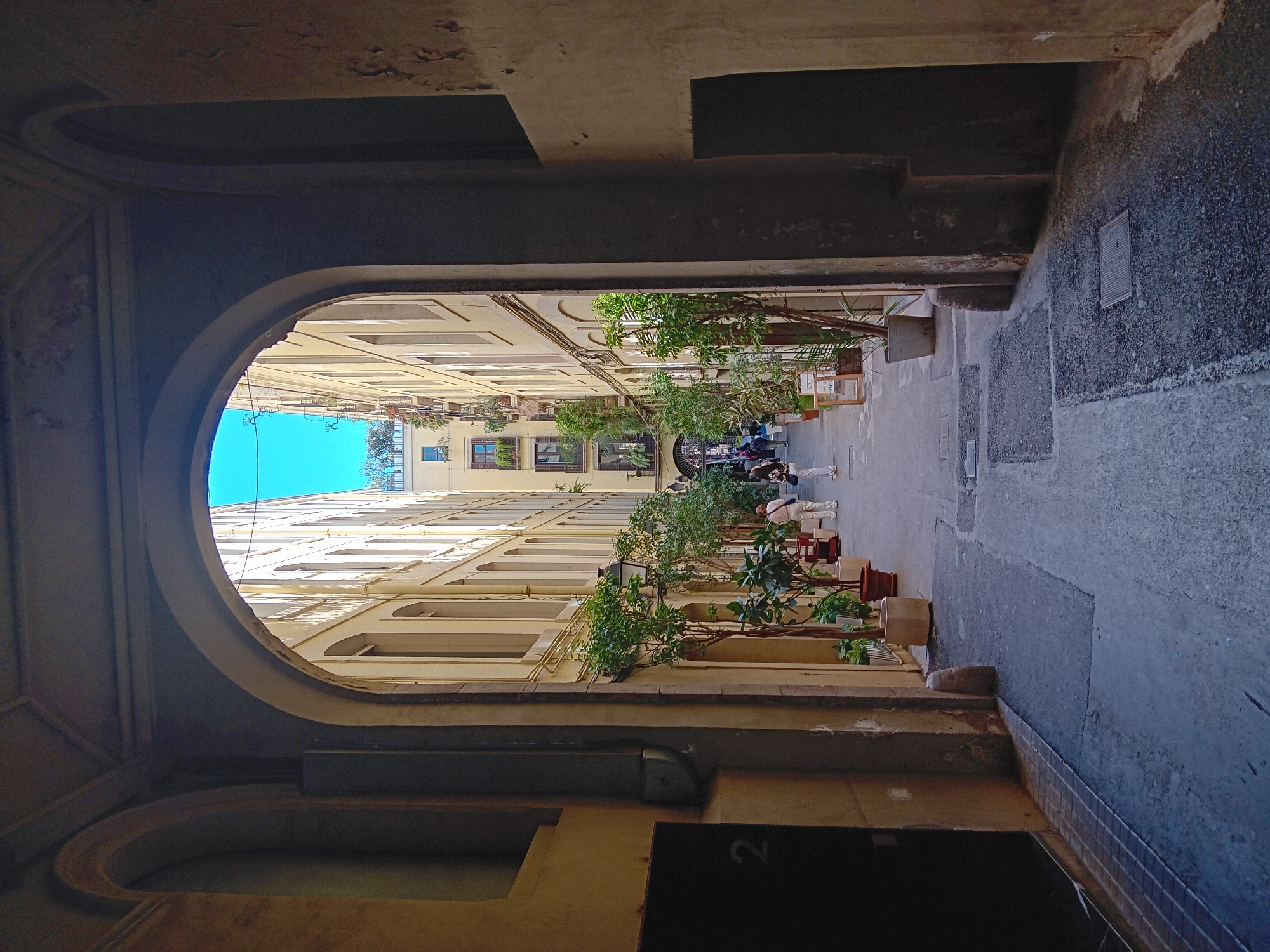 The image shows a view through an arched doorway into a narrow street in Barcelona, Spain. The perspective is from within a building, looking outwards. The archway is ornate, made of stone or concrete, with detailed moldings.
The street is lined with tall buildings, likely apartments or offices, with a repetitive pattern of windows and balconies. Plants in pots and hanging baskets adorn the balconies, adding greenery to the scene. People are present in the street, some walking and others standing near the buildings. The street is paved with a combination of stone slabs and tiles. The sky is visible through the upper part of the arch, a bright blue.