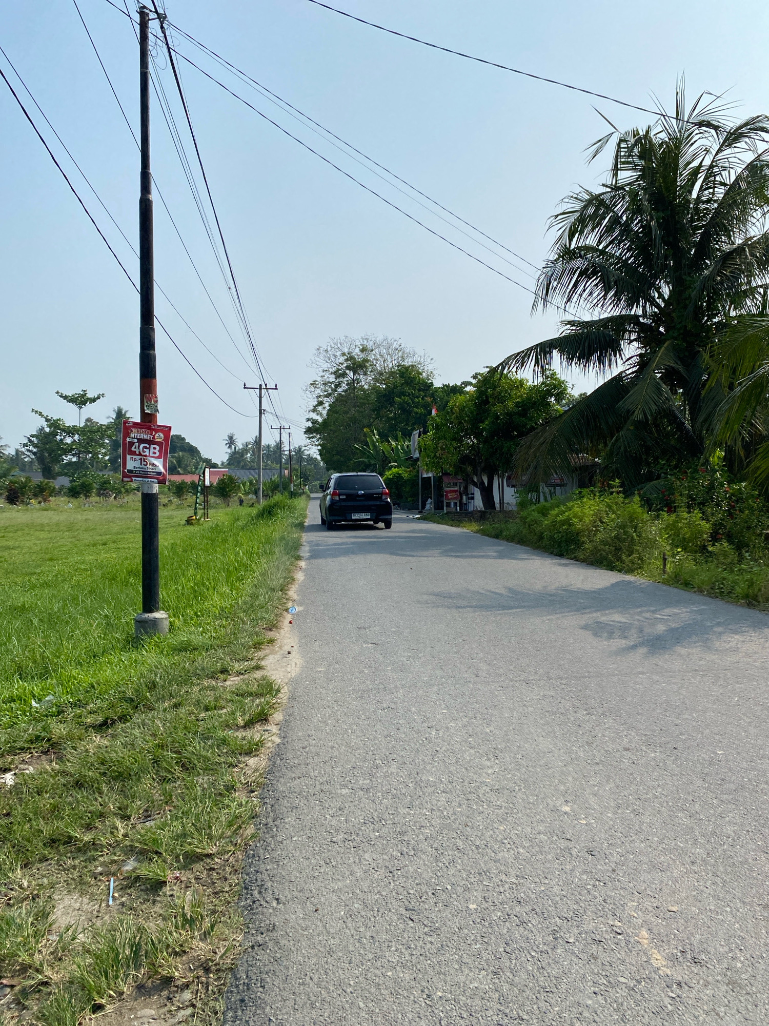 A black Toyota Avanza is driving down a paved road in Karang Gading, Indonesia. The road is flanked by a grassy field on the left and lush vegetation and palm trees on the right. A utility pole with a red and white advertisement is on the left side of the road. The sky is clear and blue.