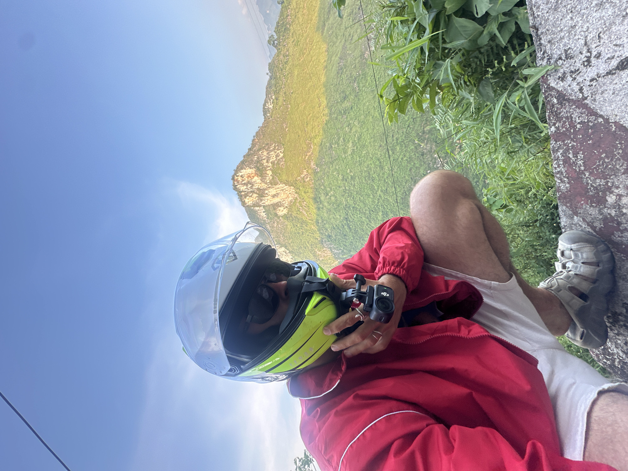 A man sits on a rock wall on the side of a road in the Đường Thượng Commune area of Vietnam. He wears a white and yellow helmet with a clear face shield, sunglasses, a red jacket with white trim, and white shorts. He holds a small black camera in his hand. He wears gray sandals. Behind him is a steep hillside covered in green vegetation. The sky is blue with some clouds.