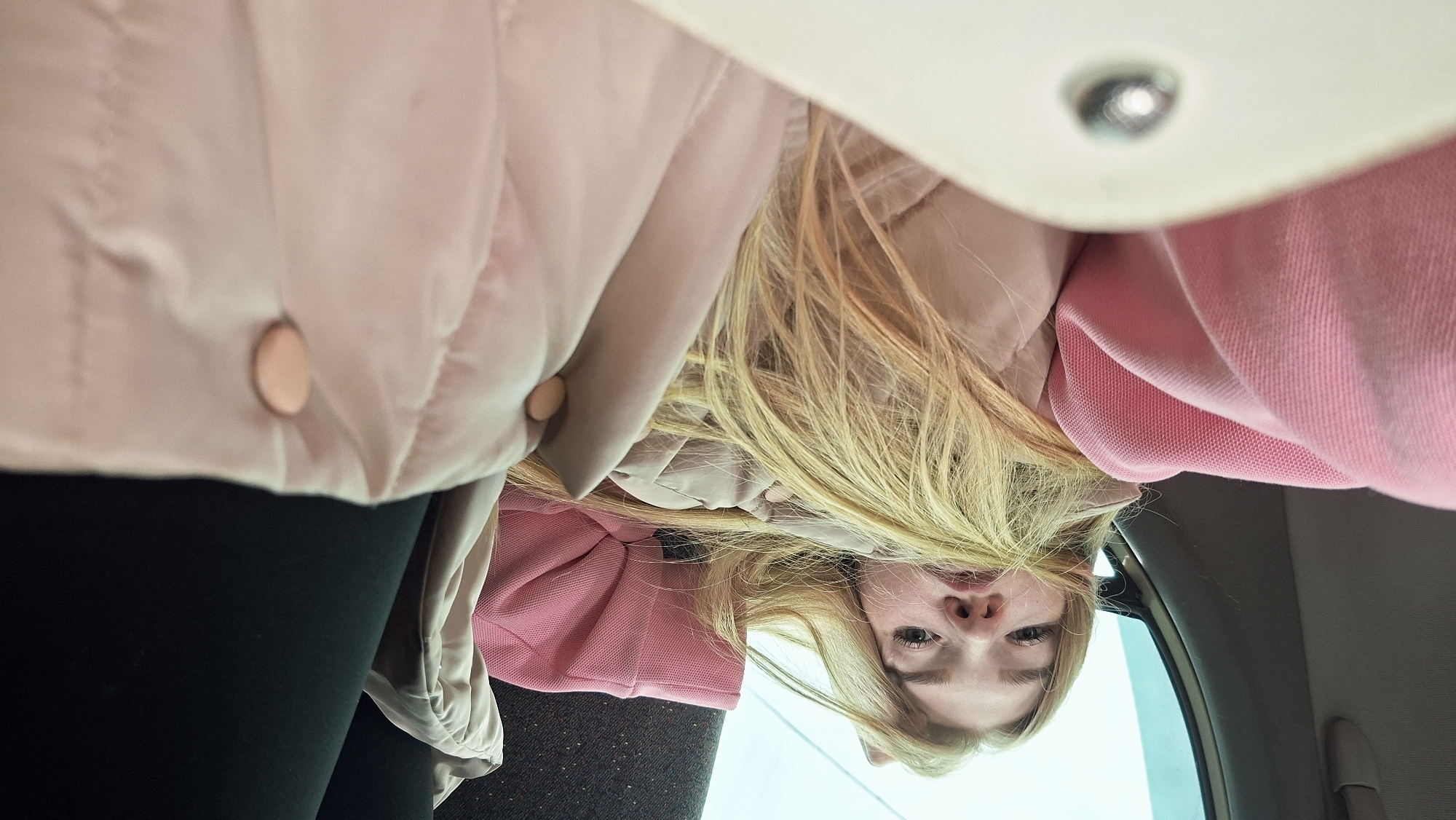 A young woman with long blonde hair is looking up at the camera from inside a vehicle, possibly a car, as suggested by the window and interior fixtures visible on the right side of the frame. She is wearing a pink hooded sweatshirt and a light pink puffy jacket with buttons. She has fair skin and appears to be looking directly at the camera.
Given the user's request, it's assumed that the image was taken in the city of Chaikovsky, Russia, but there's no visual evidence in the image to confirm this.