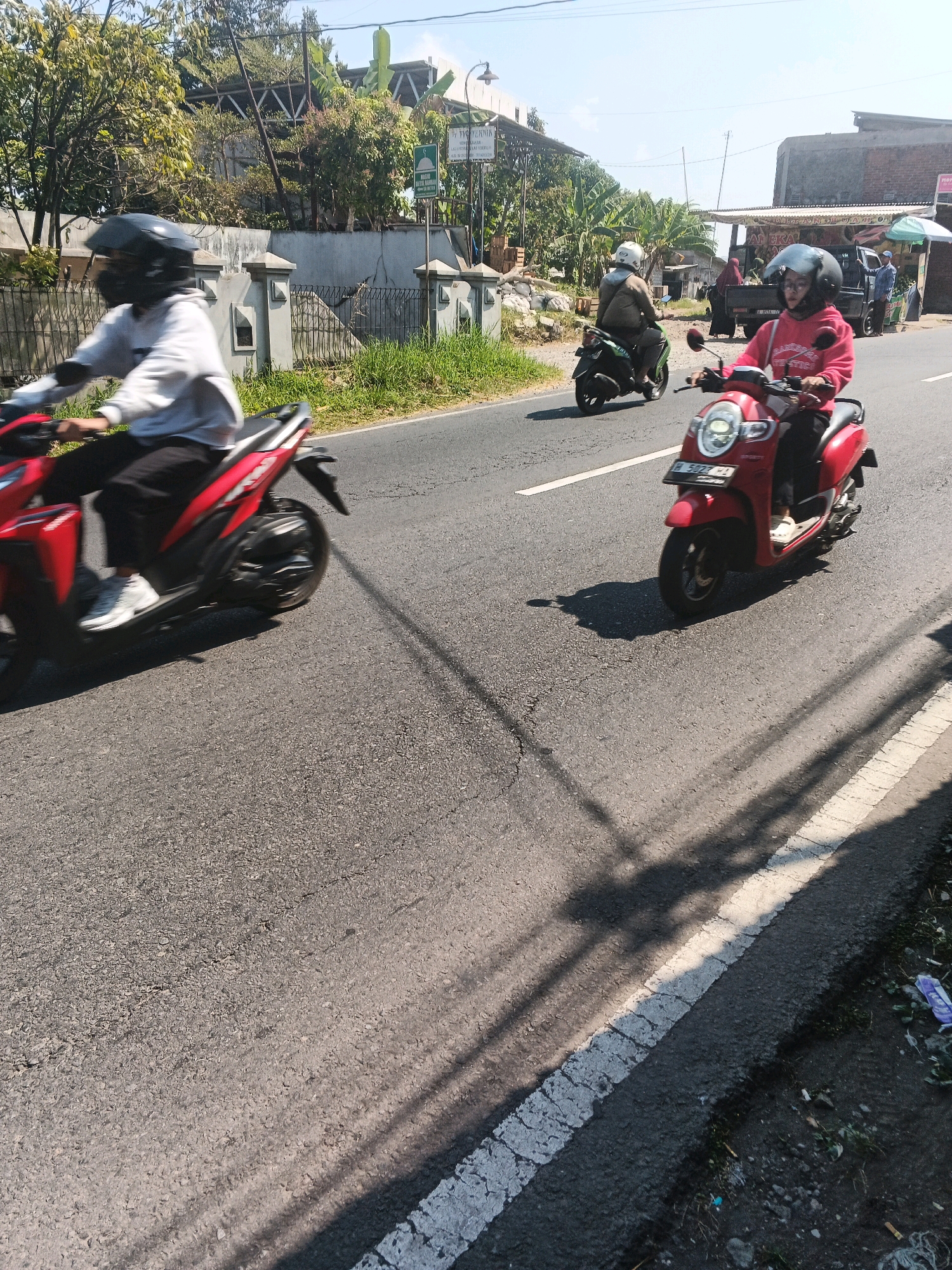 The image depicts a street scene in Bandungan, Indonesia, on a sunny day. In the foreground, a person wearing a helmet and a grey hoodie rides a red scooter. The scooter's license plate, partially visible, reads "H 5027 HL". To the left of the red scooter, another person on a red motorcycle, also wearing a helmet and a grey top, is captured in motion. Further down the road, a green motorcycle carrying a rider with a white helmet is visible. In the background, a small truck with an awning is parked, possibly a roadside stall, with several people interacting near it. The road is paved and shows signs of wear. Lush green vegetation and trees line the roadside, with buildings and structures visible in the distance. Shadows of overhead wires and trees are cast across the asphalt. The overall atmosphere suggests a typical day in a bustling Indonesian town.