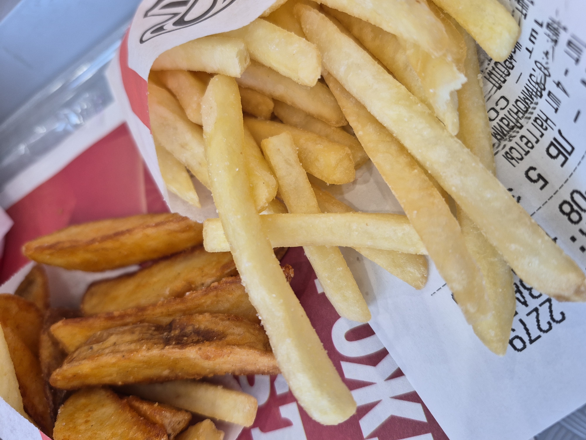 The image presents a close-up view of two types of french fries, one regular cut and one steak fry. The regular cut fries, pale yellow in color, are contained within a white paper bag or wrapper bearing red branding and lettering. A receipt or printed paper with Cyrillic text is visible alongside these fries. The steak fries, darker in color and wider in cut, are situated next to the bag of regular fries. The Cyrillic text is "ШТ - 4 ШТ Наггетсы 0.33лимонлаин ЛБ 5: 1ШТ" and the number "2279" can be seen. Tobolsk, Russia is not mentioned in the receipt so the location is undetermined.