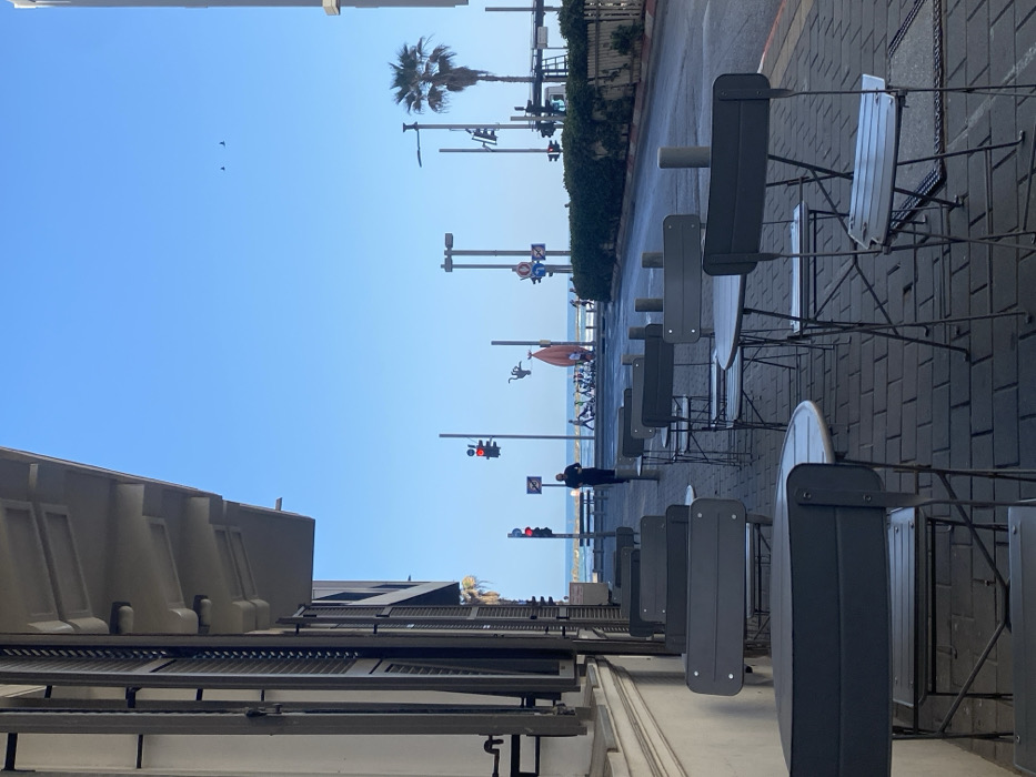 A street scene depicts empty chairs and tables lined up along a brick sidewalk. In the distance, the beach is visible, dotted with people and umbrellas. Utility poles stretch toward the sky, supporting traffic lights and various signs. A palm tree adds a touch of tropical scenery. The sky is clear and blue, with a few birds flying overhead. Buildings are visible along the street on the left.