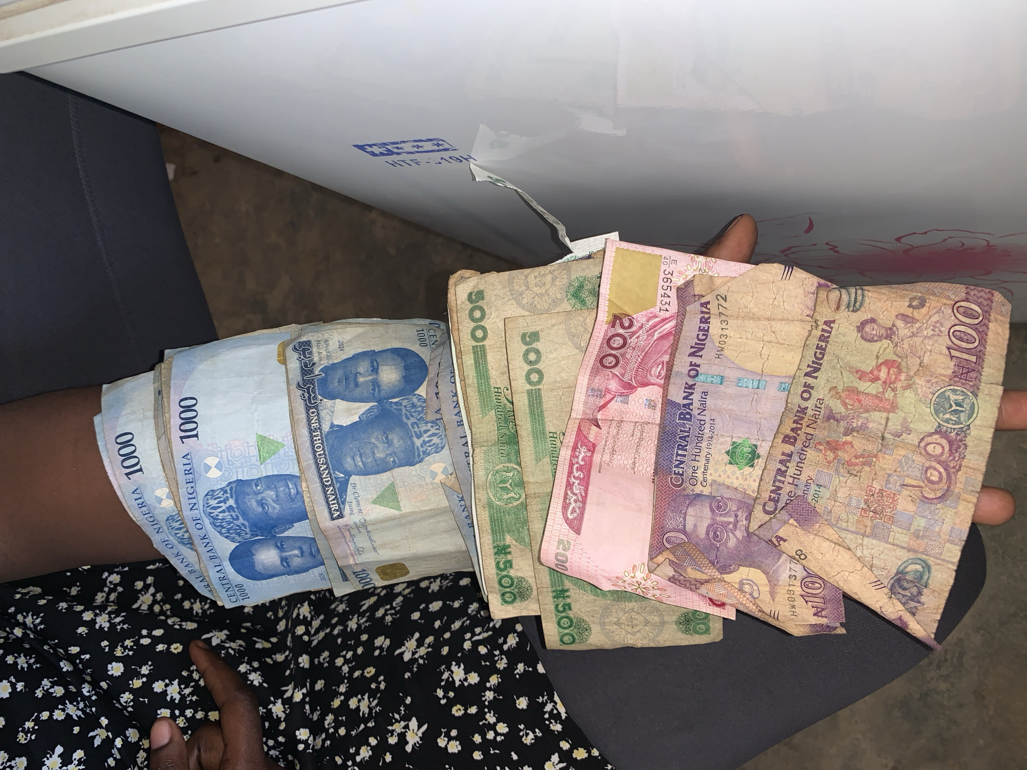 A close-up, high-angle shot captures a person's left arm and hand holding a substantial sum of Nigerian Naira banknotes, fanned out against a dark-patterned fabric, likely a dress or skirt. The money is composed of various denominations: multiple 1000 Naira notes (light blue), several 500 Naira notes (green), a 200 Naira note (pink), and several 100 Naira notes (purple/brown). The banknotes show signs of use, with some creases and wear.
The 1000 Naira notes feature portraits of Dr. Clement Isong, Alhaji Ayu, and Mai-Bomu, with "ONE THOUSAND NAIRA" clearly printed, and a date "2024". The 500 Naira notes display "Five Hundred Naira". The 100 Naira notes mention "CENTRAL BANK OF NIGERIA" and "Centenary 1914-2014", with "One Hundred Naira" and the serial numbers "HW0313772" and "HW0313710" visible on some. Other serial numbers like "H61", "40", and "365431" are also visible.
In the background, a glossy white surface, possibly a table or counter, is partially visible, with a blue stamp-like marking reading "HTF-19H". A dark, plain fabric is also seen under the money on the right. The scene is likely indoors or in a shaded area, and the activity suggests a transaction or the display of cash. The location is specified as New Stadium, Nigeria.