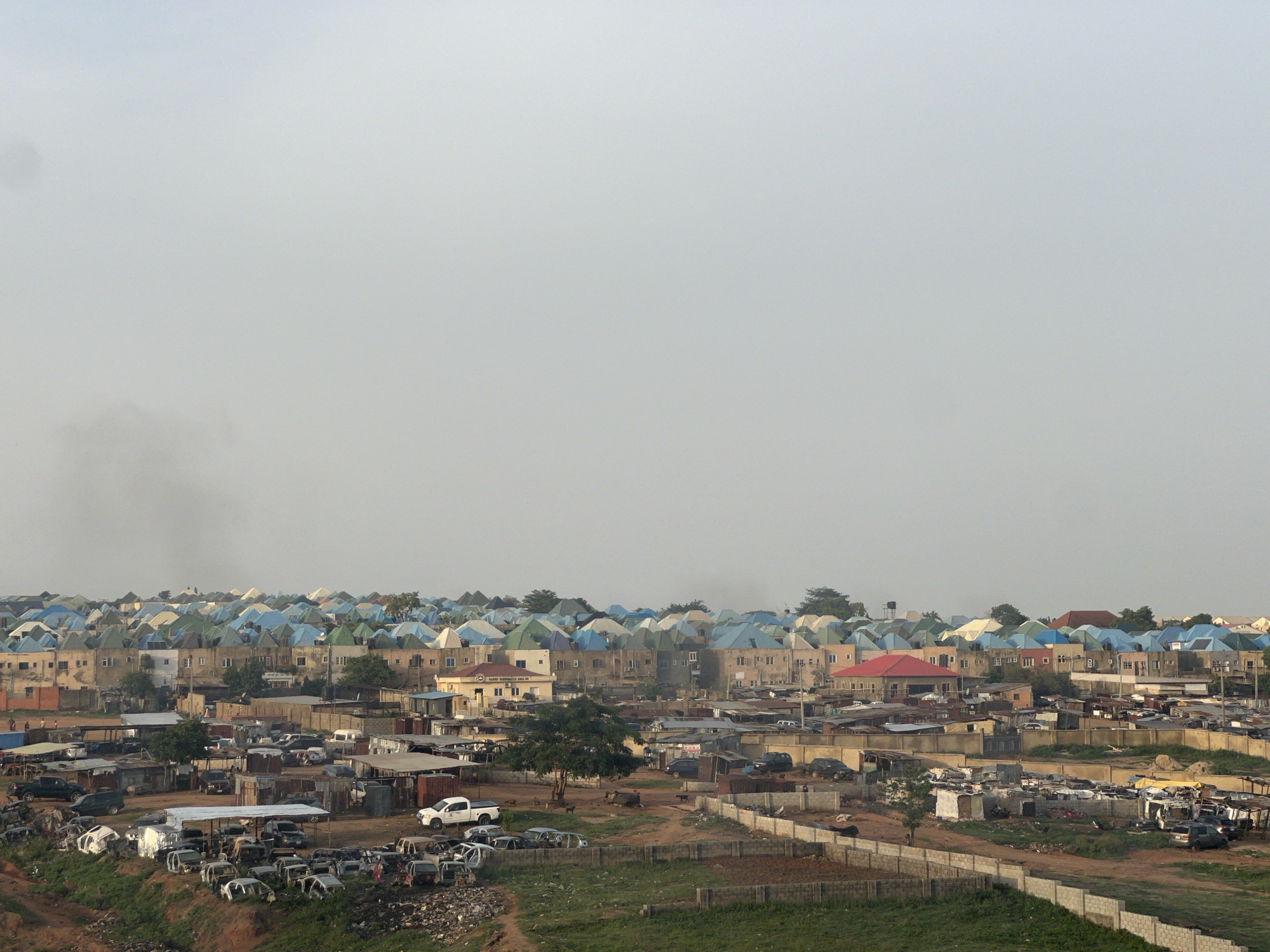 This image depicts a view of a residential area in Abuja, Nigeria. The landscape consists of numerous buildings, many with blue or green roofs, indicative of a specific architectural style. In the foreground, there is evidence of what appears to be a salvage yard or scrap area, with multiple car frames and debris scattered around. A dirt road winds through the scene, and a partially constructed or dilapidated wall is visible. There are few trees dotted throughout the area, and a green, grassy area can be seen in the immediate foreground. The sky is overcast, creating muted lighting across the scene. The overall impression is one of an urban landscape with a mix of residential areas and industrial activity, suggesting a developing or economically diverse section of Abuja.