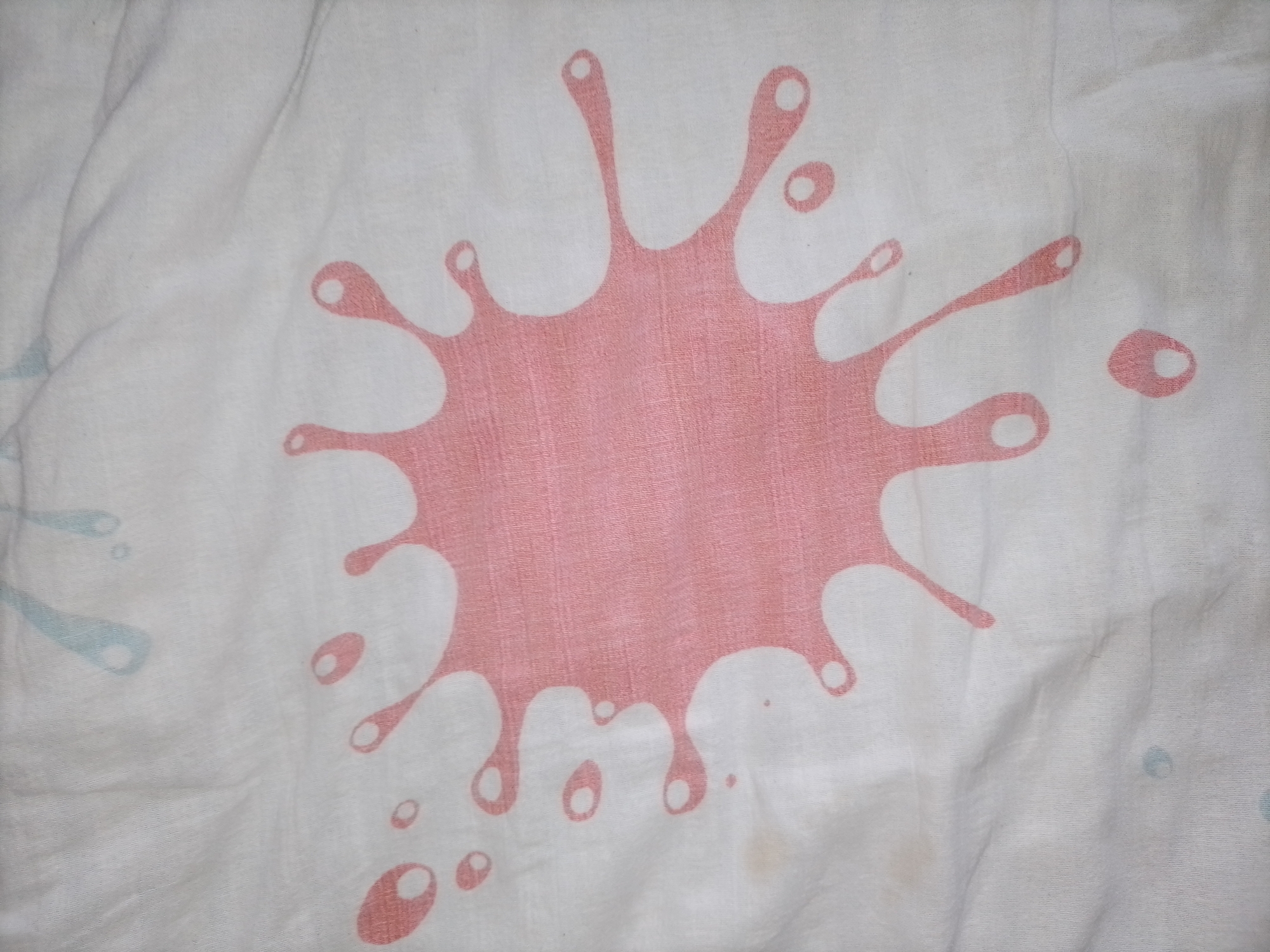The image is a close-up, top-down view of a white fabric with a pink splatter paint design. The splatter design is centrally located and appears to be the main subject. The fabric is creased and wrinkled, suggesting it is likely a piece of clothing or bedding. There are also smaller, fainter blue splatters visible on the left side of the image, and a few smaller pink splatters scattered around the main design. The lighting is somewhat dim and diffused, and there are no shadows or strong highlights, indicating an indoor setting or an overcast day. No people, text, or specific location cues are visible, other than the provided location context of Kalisz, Poland, which cannot be visually confirmed from the image itself. The overall impression is of a playful, artistic design on a textured textile.