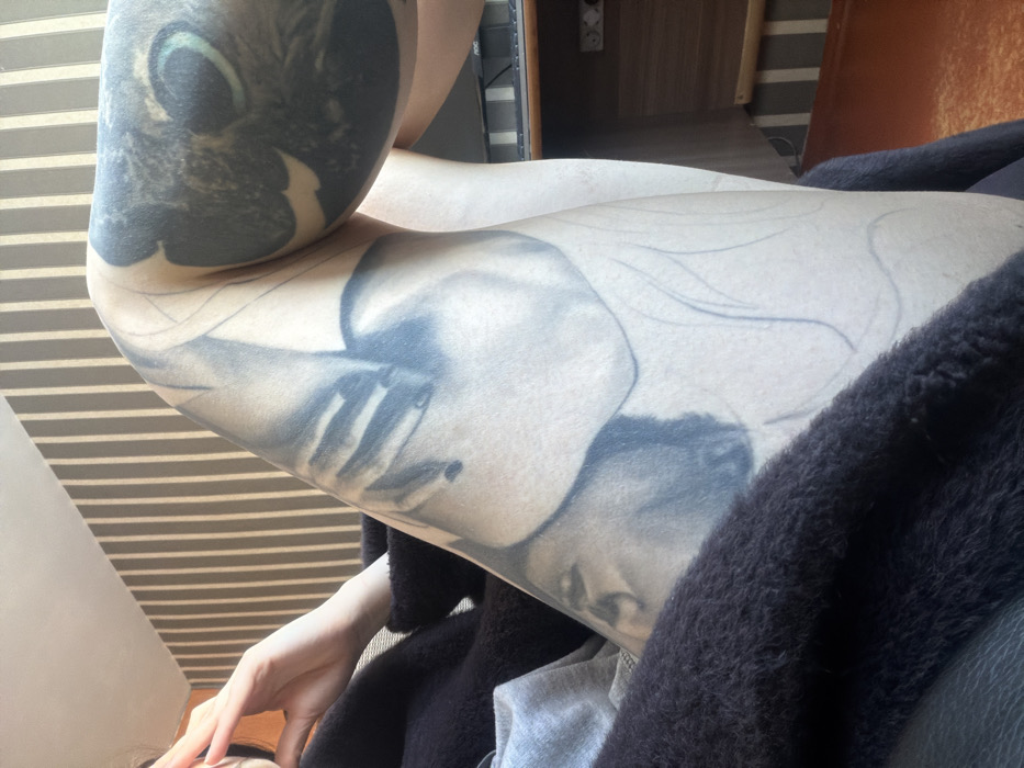 A close-up shot reveals a person's arm, heavily tattooed with a dark, surreal design featuring a face with fingers near it. Light streams through blinds casting shadows across the skin. The subject wears a dark, fluffy garment. The background shows a glimpse of a room with wooden furniture and an electrical outlet. A small portion of another person's face is visible near the bottom left corner.