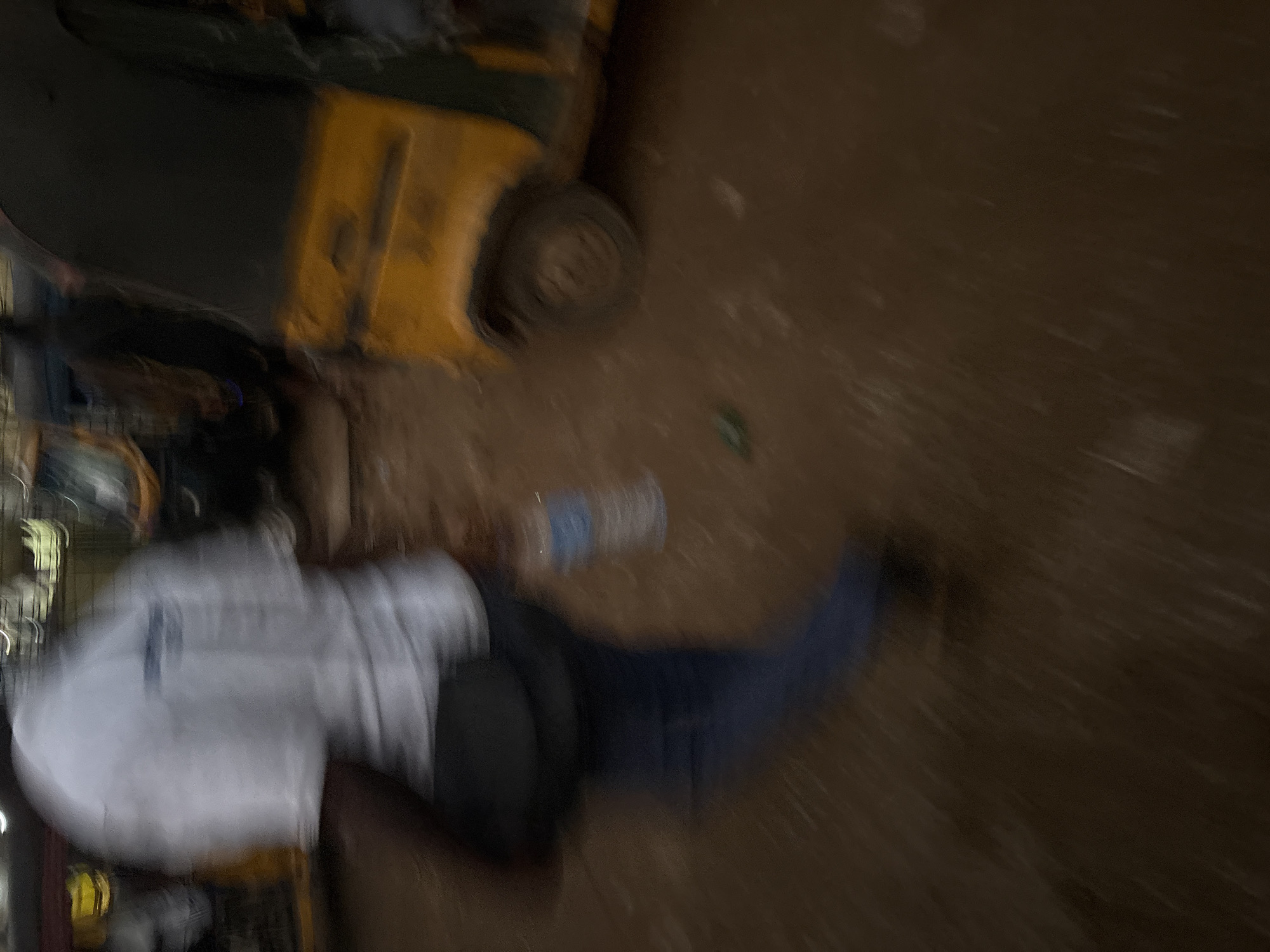The image, taken in Onitsha, Nigeria, depicts a blurred outdoor scene with a focus on a person in the foreground and a yellow auto-rickshaw in the background. The person, seen from behind, is wearing a white t-shirt and blue trousers. They are carrying a black bag slung over their shoulder. A clear plastic water bottle is visible in their hand. The ground appears to be dirt, possibly muddy, and there are other blurred vehicles or structures in the background, suggesting a street or market setting. The overall motion blur indicates movement within the scene.