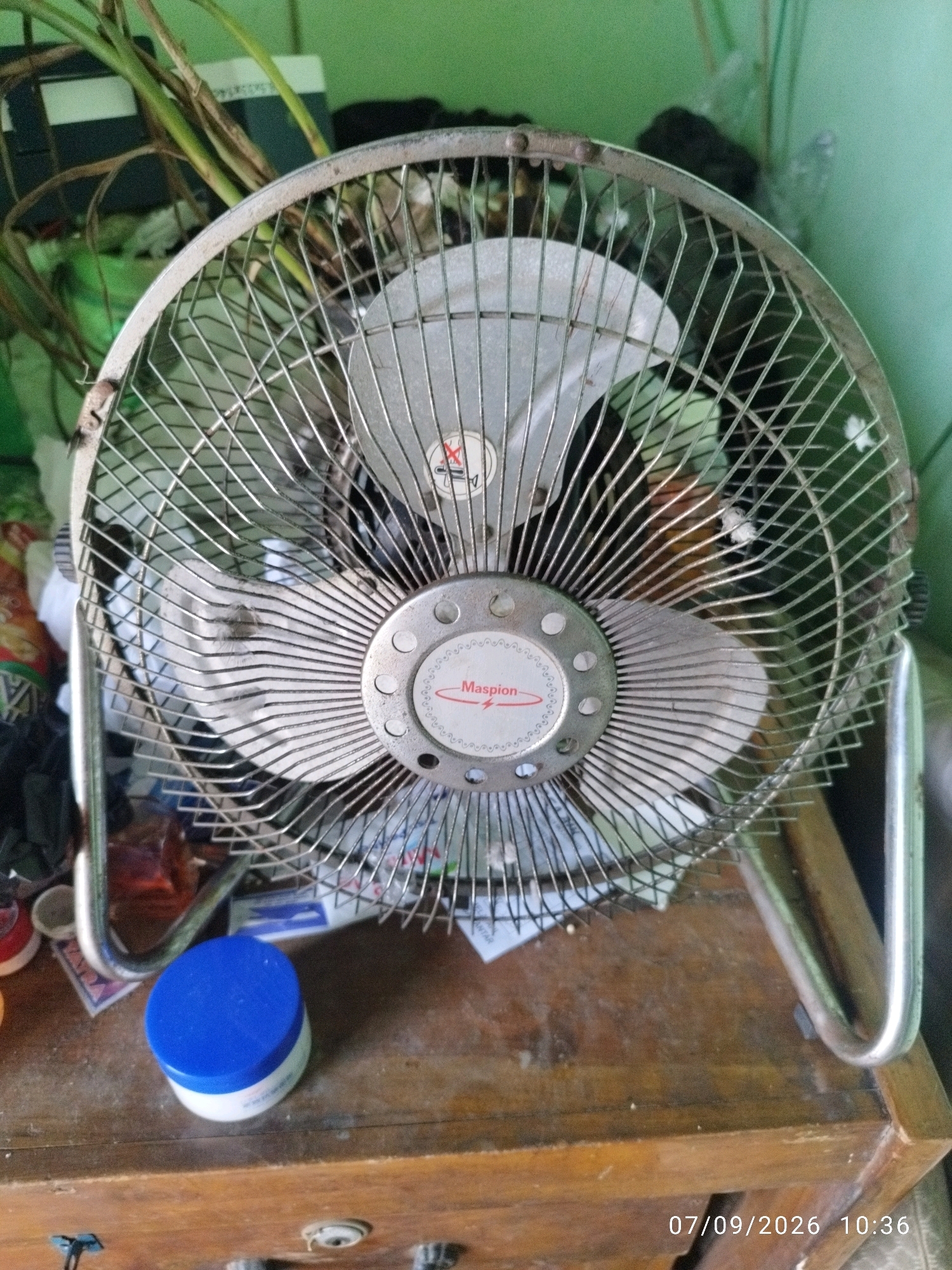 A close-up, slightly elevated shot shows a metallic desk fan with a silver grille and blades. The fan has a round, silver central housing with a red logo that reads "Maspion" and an emblem resembling a lightning bolt. The fan is positioned on a wooden surface, possibly a table or dresser, which is cluttered with various items. To the left of the fan, there is a blue-capped white container. Behind the fan, plants with long, green stalks are visible, suggesting an indoor environment. The background wall is painted a light green color. A timestamp "07/09/2026 10:36" is visible in the lower right corner of the image, indicating the date and time the photo was taken. The overall scene appears to be a domestic setting, possibly in a residential area of Prajuritkulon, Indonesia, given the timestamp format.