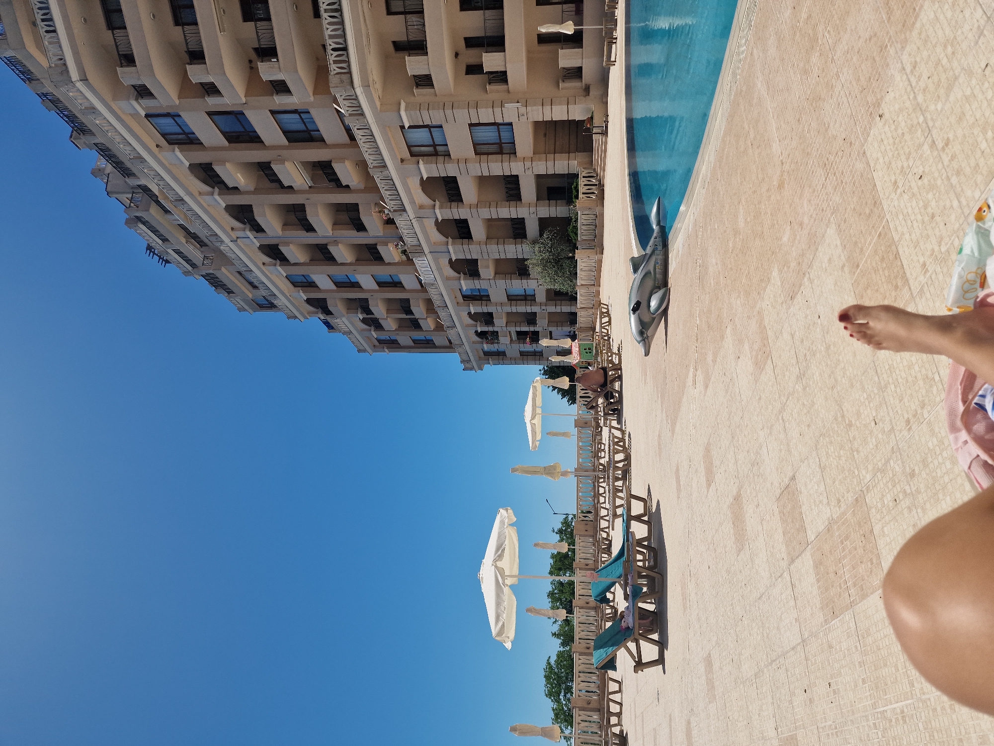 This is a daytime, eye-level, outdoor, wide shot of a resort pool area. The scene is dominated by a large, light-colored, multi-story building with many balconies and windows, set against a clear blue sky. In the foreground, a person's legs and feet are visible, indicating they are lying down. A blue swimming pool occupies the right side of the frame, with a gray inflatable dolphin floating near the edge. The pool area is paved with tan square tiles. Several empty lounge chairs are arranged on the patio, some with white umbrellas. In the background, there is a low railing, trees, and the distant sea or horizon. One person is seated in a lounge chair on the far right.