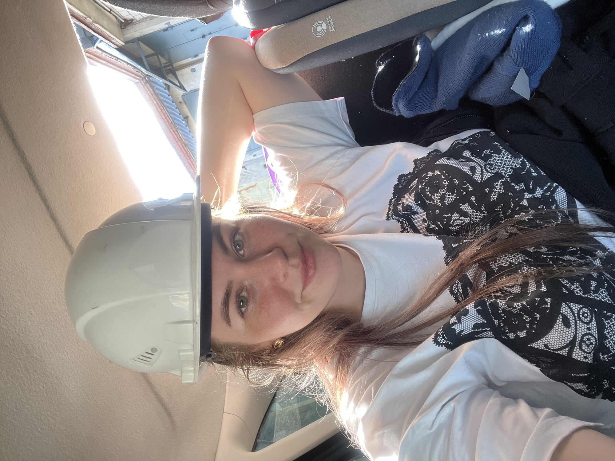 This image captures a female subject positioned within the interior of a vehicle in Chaikovsky, Russia. The subject, displaying brown hair, a fair complexion with visible freckles, and green eyes, looks towards the viewer with a slight smile. A white hard hat is placed on top of her head. She is attired in a white short-sleeved t-shirt featuring a black lace-patterned design on the chest and small gold-colored hoop earrings. Her right arm is bent, with the forearm resting on an adjacent beige and grey car seat that includes a "Side Impact Protection" label. A rolled-up blue fabric item and other dark garments are visible on the seat next to her. Strong sunlight enters the vehicle from the upper right, illuminating the subject's hair and the car's beige headliner. Through the vehicle's window, an exterior building is partially observed, characterized by reddish-brown and blue corrugated roofing, light-colored walls, and dark rectangular features, indicative of an industrial or garage environment.