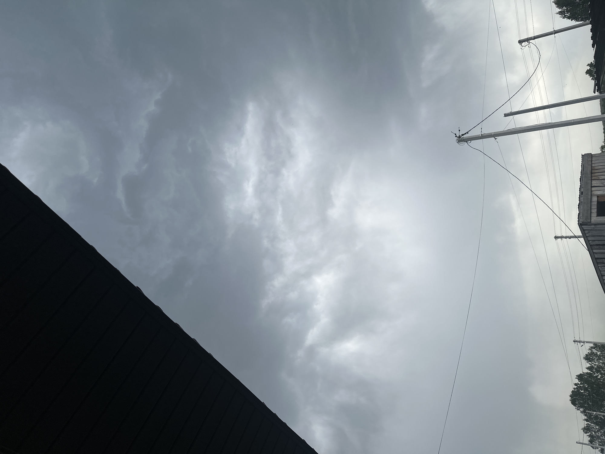The image shows a low-angle view of an overcast sky with dark gray clouds. In the foreground, the corner of a dark roof is visible. On the right side, utility poles and power lines are present. The surrounding environment includes trees and a weathered building. The location, Kharnavy, Russia, is not discernible from the image alone.
