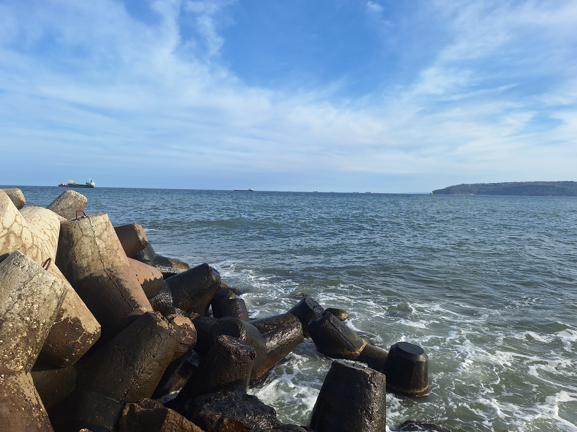The image depicts a coastline scene in Varna, Bulgaria, dominated by concrete tetrapods. These structures are clustered together in the foreground, with some partially submerged in the water. The sea stretches out behind the tetrapods. There are ships on the horizon. A landmass is visible on the right side of the horizon. The sky is blue with scattered clouds. The photograph appears to have been taken during daylight hours.