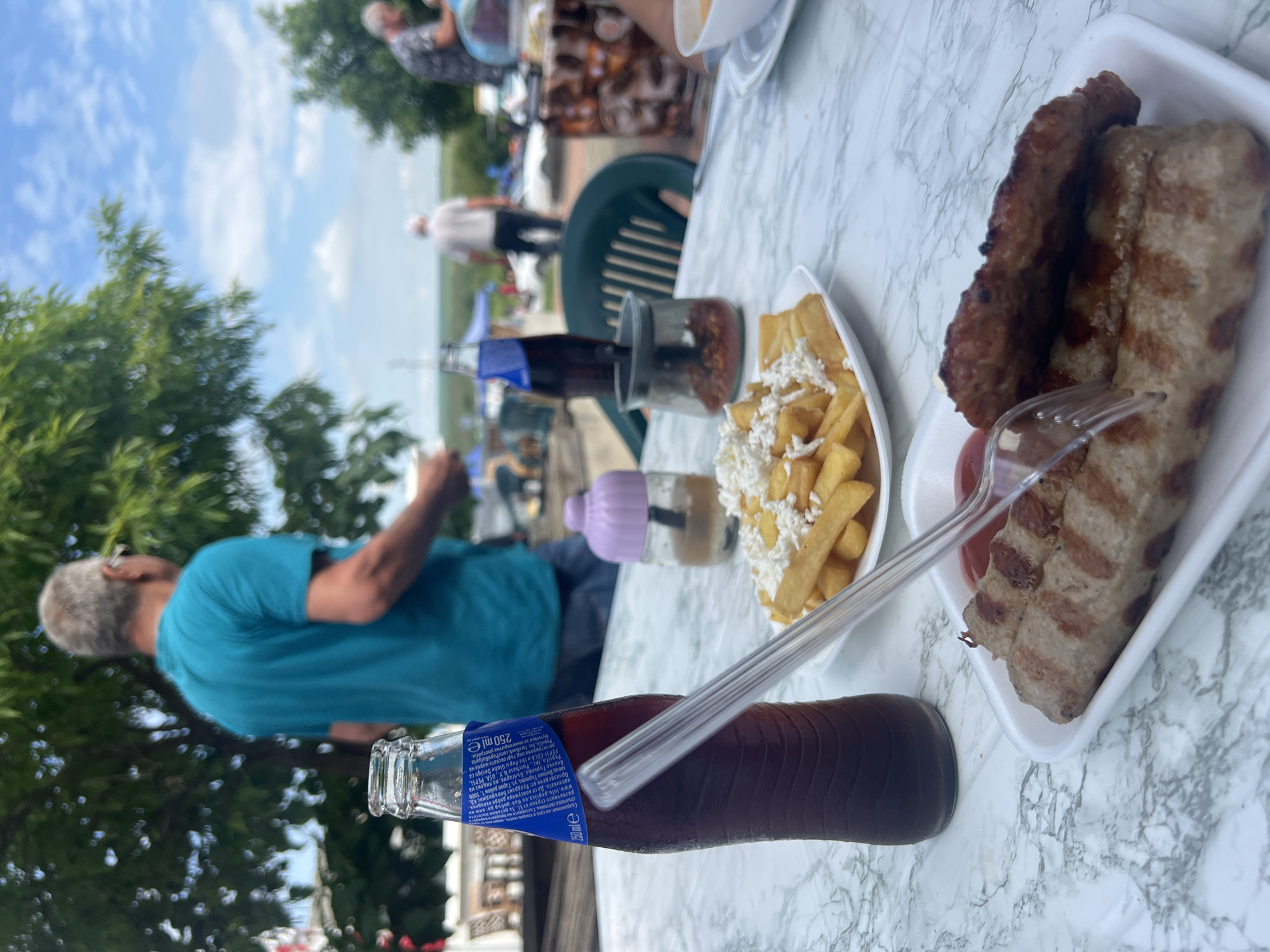 The image depicts an outdoor dining scene in Hitrino, Bulgaria. In the foreground, a white marble-patterned table holds a clear glass bottle of dark soda, 250ml in volume, with a blue label featuring Cyrillic script and condensation on its surface, containing a transparent plastic straw. Beside it is a white rectangular foam plate with two grilled oblong meat items, one pierced by a transparent plastic fork, and a small amount of red condiment (ketchup) visible. Adjacent is a white round plate containing french fries topped with crumbled white cheese. To the left, a small, light purple lidded container holds a translucent liquid. To the right, a small glass jar contains red flakes, and a portion of a dark green slatted plastic chair is visible. In the midground, out of focus, a man in a teal t-shirt and glasses stands facing away from the camera, beneath green foliage. The background features a bright sky with scattered white clouds, more blurred figures, trees, and distant structures including a white vehicle and tents, suggesting an open public area.