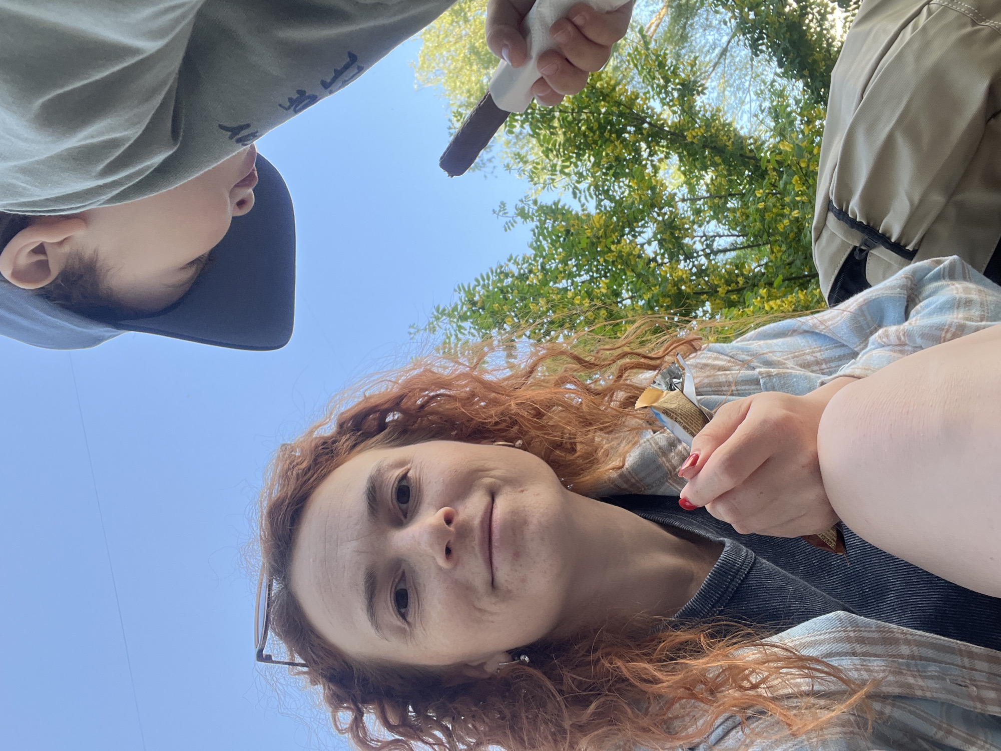 The image is a low-angle shot looking up at three people and the sky. The main subject is a woman with long, curly red hair. She is wearing glasses and a blue and tan plaid shirt over a dark blue shirt. She is looking directly at the camera and holding a chocolate bar in her hand. Above her is a child wearing a gray hooded shirt and a navy blue hat. Their face is partially visible. To the right of the child, someone is holding another chocolate bar towards the camera. In the background, there are green leafy trees and a clear blue sky. The location is the city of Chaikovsky, Russia.
