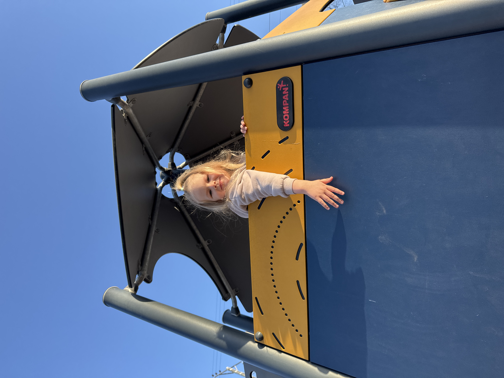 A young girl is peering out from a blue and yellow play structure at a playground. The structure has a metal canopy overhead. The logo "KOMPAN!" is visible on a yellow panel. The background consists of a clear blue sky. The location is Perm, Russia.