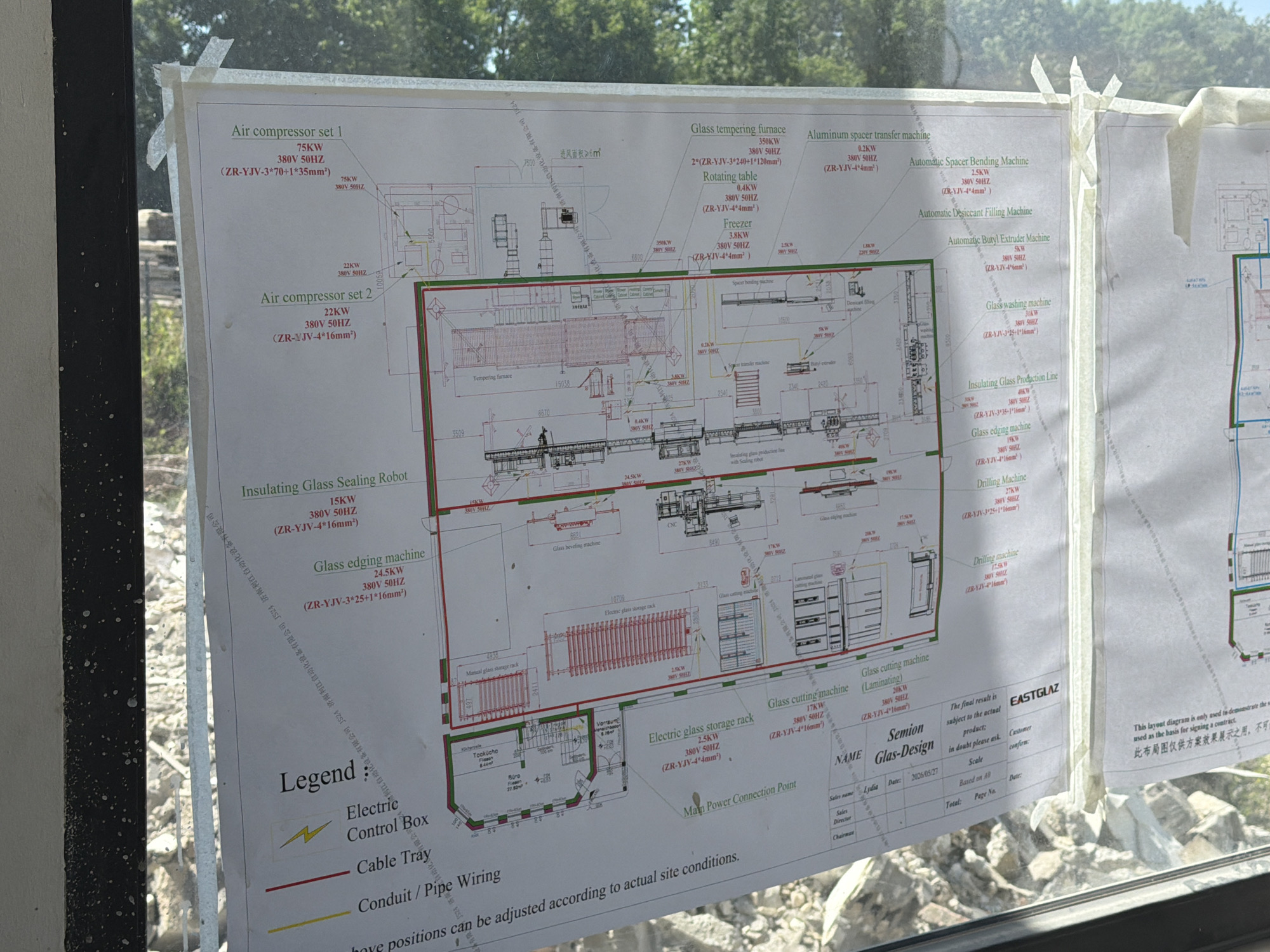 This is a medium shot of a technical diagram displayed on a large sheet of paper, seemingly taped to a window or glass surface, as evidenced by reflections and the blurry background of trees and sky. The diagram itself is a complex blueprint for a manufacturing facility, likely for glass processing.
The diagram is densely packed with technical information, including labels for various machines and their specifications (e.g., "Air compressor set 1," "Glass tempering furnace," "Insulating Glass Sealing Robot," "Glass edging machine," "Drilling Machine," "Electric glass storage rack," and "Glass cutting machine"). Electrical specifications like KW ratings, voltage (380V), and frequency (50HZ), along with cable types and sizes (e.g., "ZR-YJV-3*70+1*35mm²"), are prominently featured.
A "Legend" section at the bottom left explains symbols for "Electric Control Box," "Cable Tray," and "Conduit / Pipe Wiring." There are also sections for project details, including "NAME," "Semion Glas-Design," and fields for "Sales name," "Sales Director," and "Chairman," with a date "2026/05/27" visible. Company names like "EASTGLAZ" and "济南利江自动化设备有限公司" are also present.
The overall impression is one of a detailed technical plan or proposal. The presence of tape suggests it's a temporary installation, possibly for review or client presentation. The lighting indicates it's daytime, with natural light filtering through the paper and the background. The background is out of focus, showing a natural environment with trees and foliage, suggesting the facility might be located in a semi-rural or industrial area. No people are visible in the image.