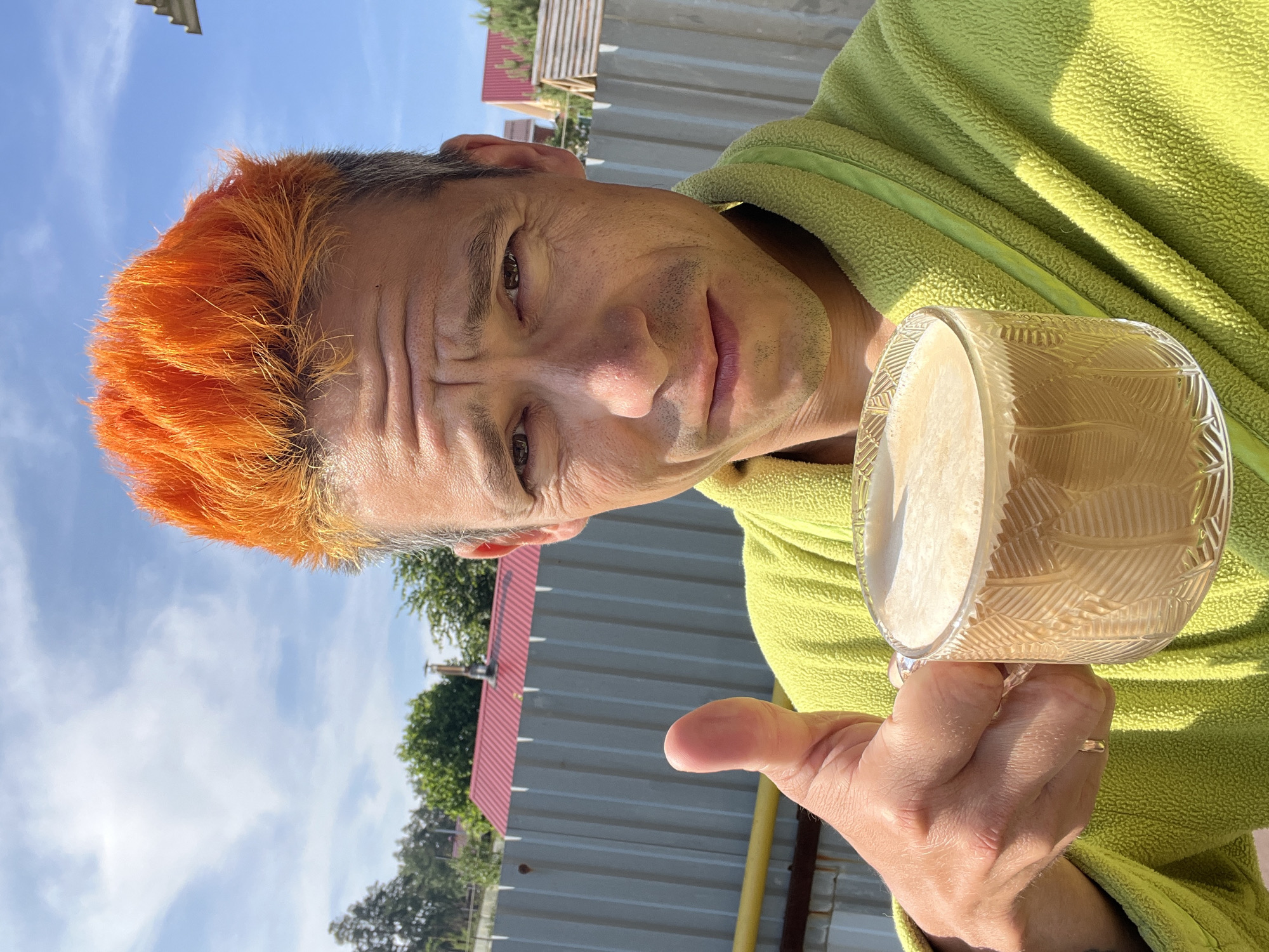 A man with bright orange hair stands outdoors, giving a thumbs-up while holding a clear glass mug of coffee. He is wearing a light green bathrobe. In the background, a corrugated metal fence and trees are visible under a blue sky with some clouds. The image appears to be taken in Chaikovsky, Russia, based on the provided information.