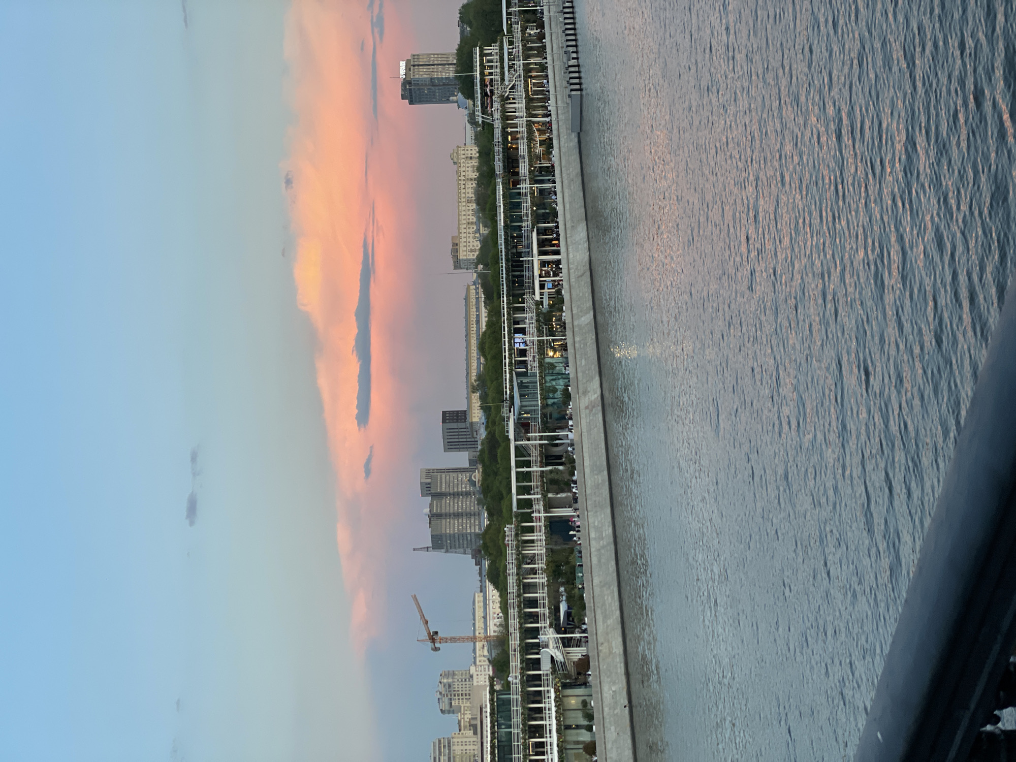 The image shows a cityscape at dusk. In the foreground is a body of water, its surface reflecting the colors of the sky. Adjacent to the water is a promenade lined with structures, possibly restaurants or cafes, with visible architectural details. Buildings of varying heights and designs line the skyline. A construction crane is visible among the buildings. The sky features hues of blue, pink, and orange, suggesting a sunset. A dark railing is in the immediate foreground.