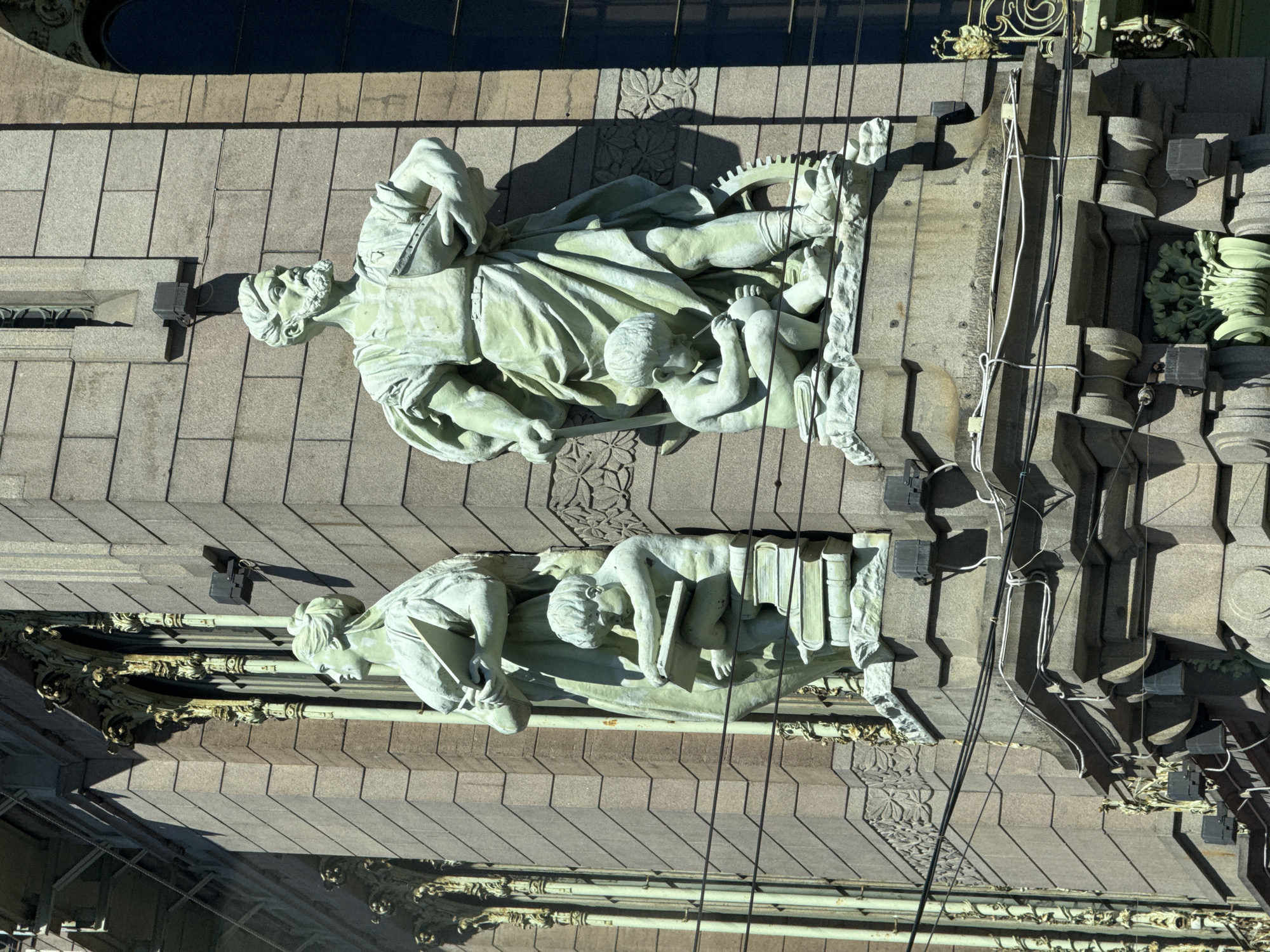 This media file displays an aerial view of architectural sculptures on the facade of a building, likely in Saint Petersburg, Russia. The primary subjects are two distinct sculptures of human figures, rendered in a pale, possibly patinated, material.
The upper sculpture depicts an adult figure, appearing to be male with a beard, draped in flowing robes. This figure is shown in a dynamic pose, with one arm outstretched and holding what appears to be a staff or rod. The sculpture also includes a smaller, child-like figure nestled within the folds of the adult's garment, reaching out towards the adult's hand. A gear or cogwheel is visible behind the adult's legs, hinting at a theme of industry or progress.
Below this, a second sculpture features two figures. One appears to be an adult, possibly female, leaning over a book or tablet. Another younger figure is positioned in front, also engaged with a book. Several books are depicted stacked beside the younger figure, suggesting a theme of learning or scholarship. Both sculptures are detailed with drapery and facial features, and appear to be integrated into the building's decorative elements, which include floral motifs and architectural molding.
The lighting suggests it is daytime, with strong shadows cast by the sculptures and architectural features, indicating a bright, sunny day. The scene is devoid of any overtly discernible human activity beyond what is depicted in the sculptures themselves. The setting is clearly an exterior architectural facade, with stone cladding and ornate details typical of historical European architecture. No text is visible in the image.