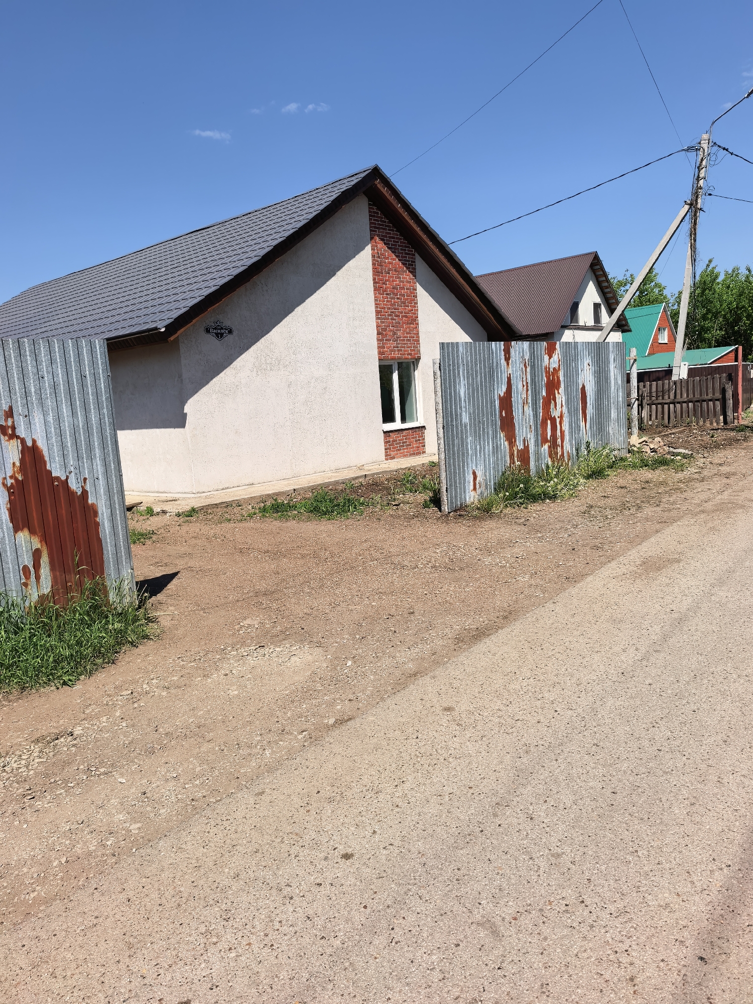 The image shows a residential area in Sterlitamak, Russia. A one-story house with a gray roof and beige stucco walls is visible. A section of the house is constructed of red brick. The house features a small window on the brick section and a sign with Cyrillic lettering on the stucco wall. A corrugated metal fence stands in front of the house. The fence exhibits rust stains. Behind the house, additional houses with brown and green roofs are partially visible. A utility pole with electrical wires is also visible. The foreground is dominated by a dirt road with grass growing along the edges. The sky is clear and blue.