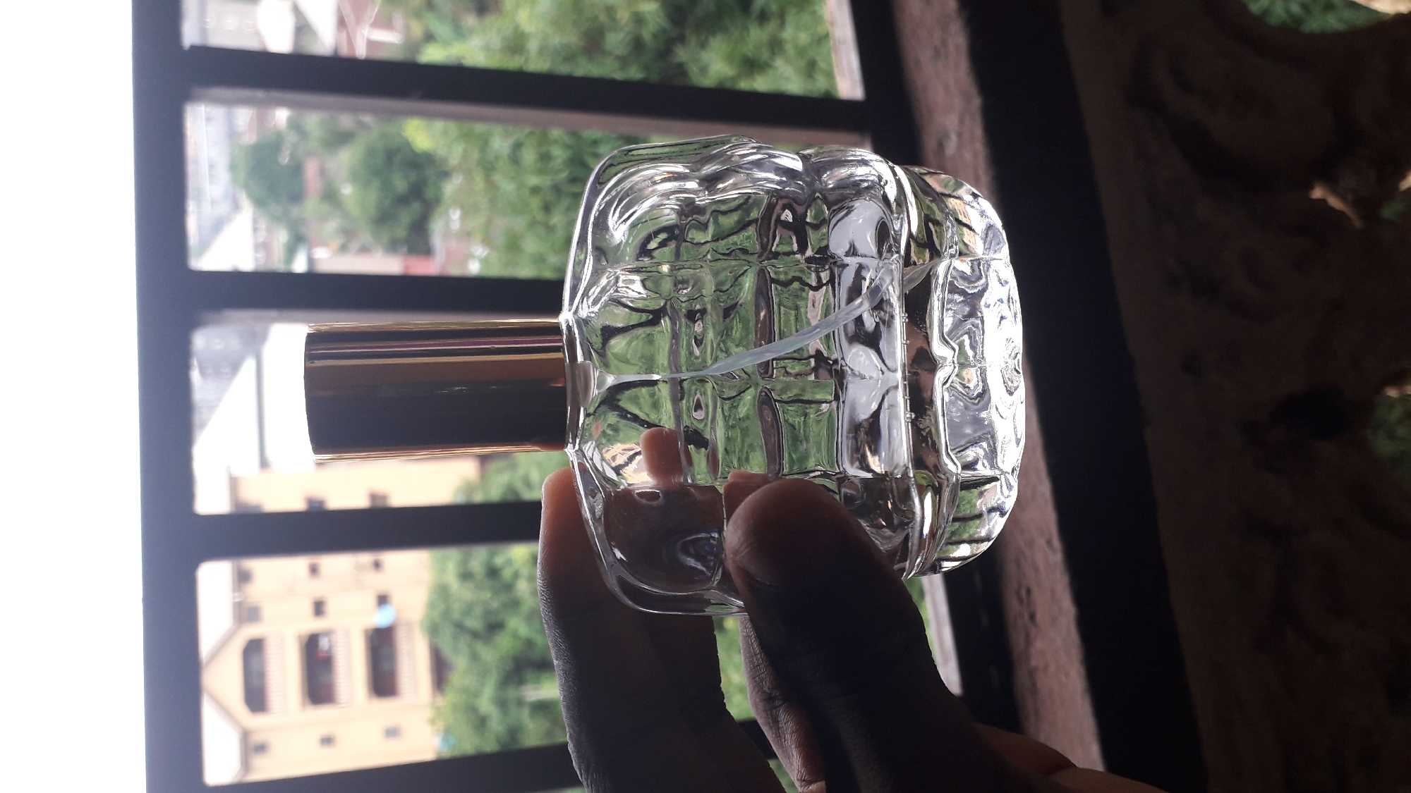 A person's dark-skinned hand holds a clear glass perfume bottle with a gold-colored cylindrical cap. The bottle has a multifaceted, geometric design. The background is a window with dark metal bars, through which buildings and lush green trees are visible. The scene appears to be indoors, based on the window frame and the presence of what seems to be carved wood paneling on the right side of the frame. The location is identified as Nkpor, Nigeria.