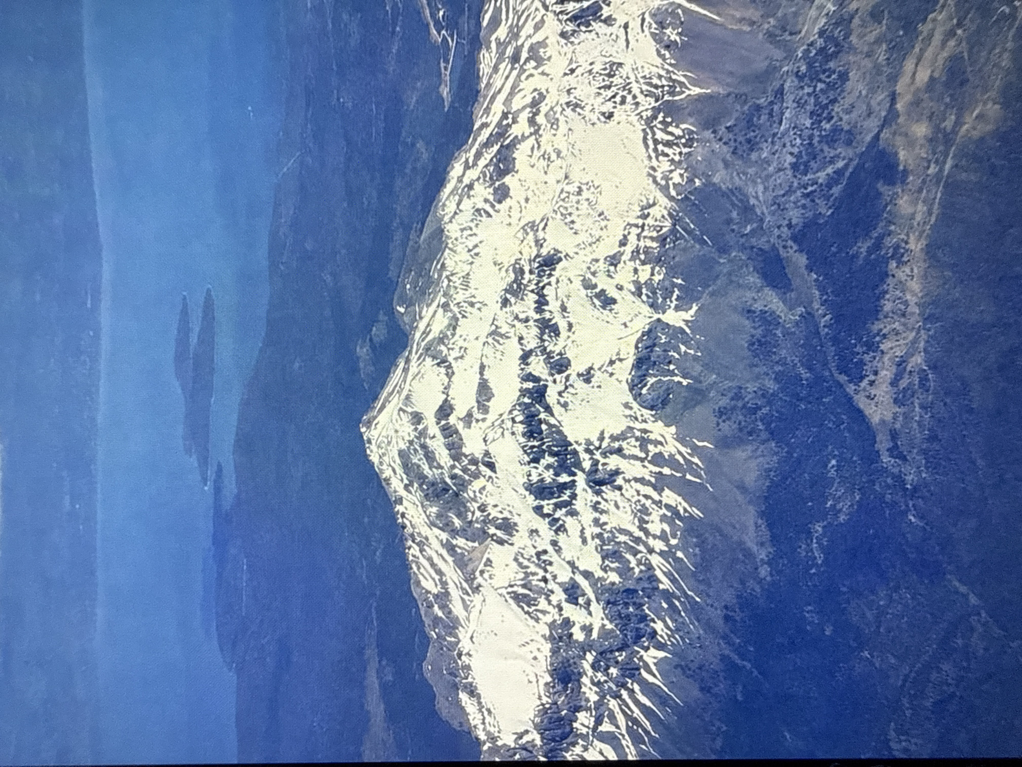 This is a rotated aerial photograph showing a snow-capped mountain range adjacent to a large body of water. The mountain peaks are jagged and dramatic, with stark contrasts between the snow and the dark rock. Patches of snow are scattered across the slopes, with some areas appearing to be glaciers or permanent snowfields. The water is a deep blue and appears calm, with a hint of a distant shoreline or islands. The lighting suggests it is daytime, with shadows indicating the sun's position. The scene conveys a sense of vastness and natural beauty, with no human presence visible. The rotation of the image suggests it may have been captured from an unusual angle, possibly from an aircraft or drone. No text is visible in the image. The context of Mykolaiv, Ukraine, is noted, but there are no specific visual cues within the image to confirm this location.