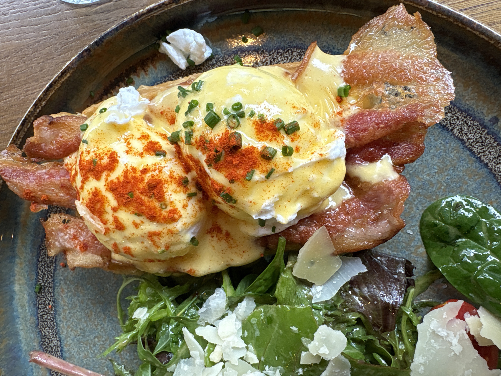 A high-angle, close-up shot depicts a plate of Eggs Benedict with bacon and salad. Two poached eggs are arranged on top of toasted bread and bacon, covered with hollandaise sauce, garnished with green chives and paprika. Crispy bacon strips are positioned around the eggs. A mixed green salad with cheese shavings occupies the lower portion of the plate. The dish is served on a textured plate with blue, brown, and gold accents. The setting is indoors, likely a restaurant. There is no evidence to suggest that the image was taken in Sofia, Bulgaria.