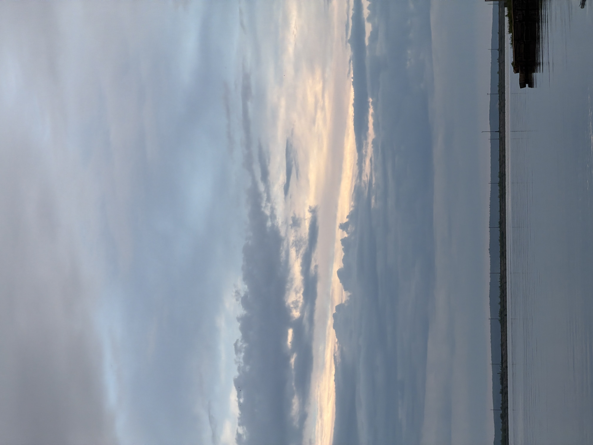 The image depicts a body of water, likely a lake or river, with a cloudy sky overhead. The water is calm and reflects the sky. A distant shoreline is visible with what appear to be utility poles or other vertical structures along it. A small section of what appears to be a dock or other wooden structure is visible along the top edge of the image on the water. There are no people visible in the image. Based on the prompt information, the location is the city of Chaikovsky, Russia.
