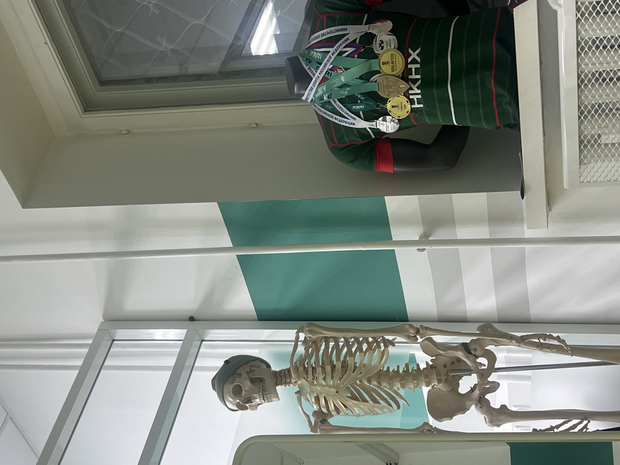 The image, taken in Kazan, Russia, displays an indoor scene featuring a mannequin and a human skeleton model within what appears to be a museum, educational facility, or sports hall. The background wall is painted with alternating white and teal-green horizontal stripes.
On the right side of the frame, a dark-skinned mannequin stands, wearing a dark green sports jersey. The jersey is adorned with red and white vertical stripes and the prominent white Cyrillic text "HKHX" on the chest. Several medals of gold, silver, and bronze colors are draped around the mannequin's neck. Visible Russian text on the medal ribbons and medals includes "МИНИСТЕРСТВО СПОРТА РОССИЙСКОЙ ФЕДЕРАЦИИ" (Ministry of Sport of the Russian Federation), "PARI", "1 МЕСТО" (1st Place), "ПОБЕДИТЕЛЬ" (Winner), and "МЕСТО" (Place).
On the left, a full human skeleton model is displayed upright inside a glass case. The skeleton's skull is covered with a dark, beanie-like cap. A skylight or window is visible towards the top of the scene, allowing for some natural light. No specific activity is occurring; the scene is a static display commemorating sports achievements and showcasing anatomy.