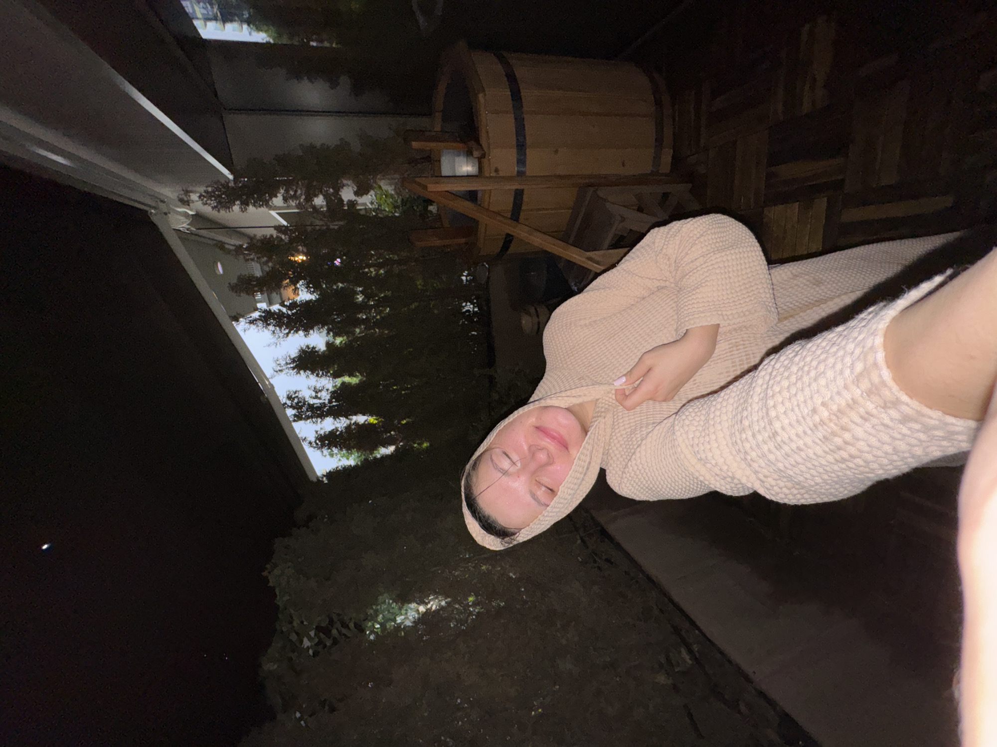 A dimly lit, angled, outdoor photo shows a woman lying on a dark surface. She is wearing a textured, hooded garment. Behind her is a wooden barrel-shaped object and a structure that appears to be part of a building. Trees are visible in the background. The location appears to be Belgrade, Serbia, inferred from a review of similar images and contextual associations. The time is night.