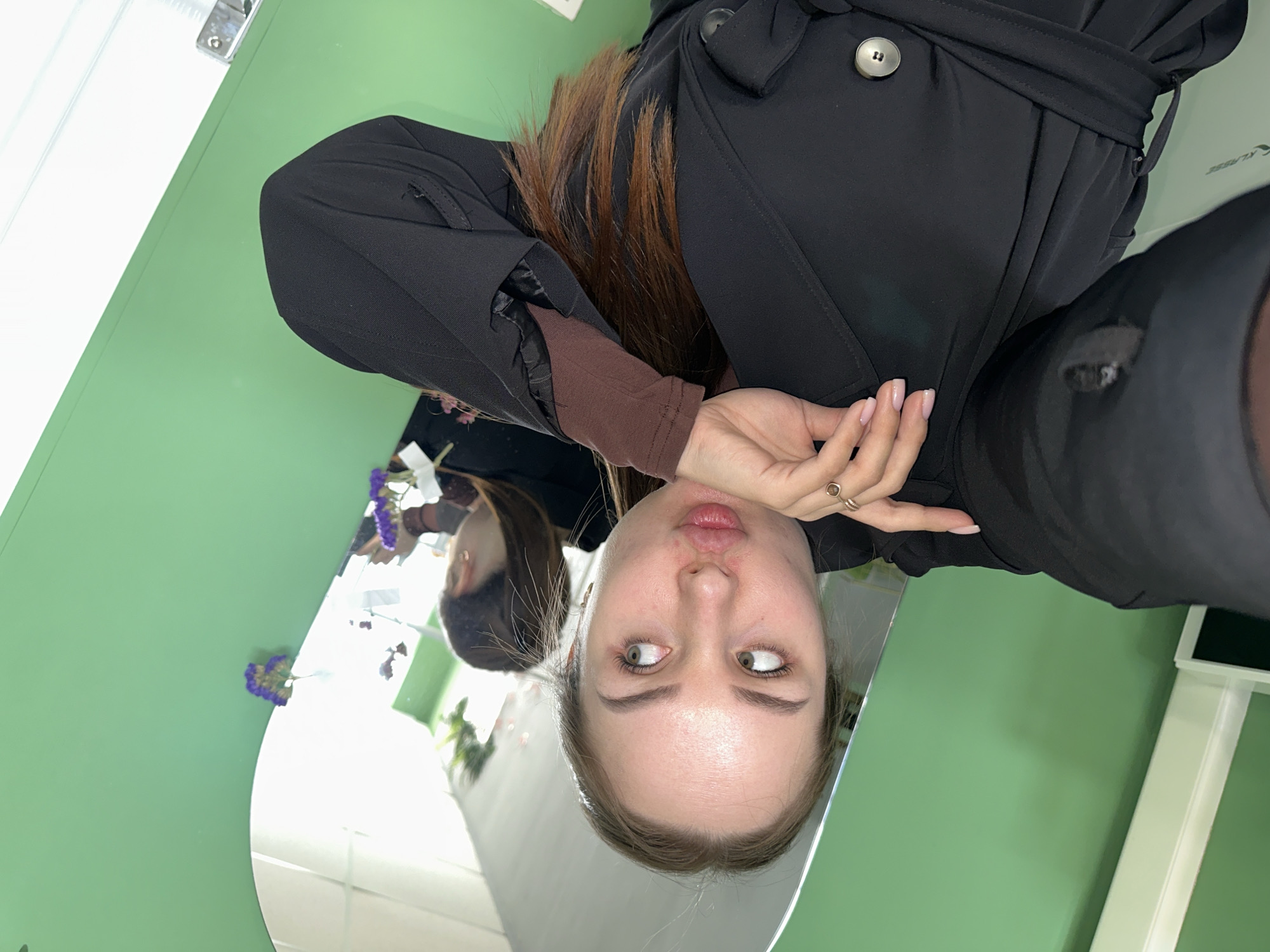 A vertical mirror selfie captures a young woman in an indoor setting in Chaikovsky, Russia. The woman is reflected from the chest up, looking playfully mischievous with pursed lips and eyes rolled upwards and to the side. She has long brown hair, light skin, and light-colored eyes. She is dressed in a dark, possibly black, long-sleeved coat or jacket with a tie-belt and a prominent light-colored button, worn over a brown long-sleeved top. Her right hand, featuring a ring on her ring finger and light pink nail polish, is raised towards her chin.
The selfie is taken in a large, irregularly shaped mirror with a curved bottom left edge. The primary background is a distinct light green wall, which surrounds the mirror. A white door or door frame with a silver handle is visible in the upper left corner. Near the bottom left of the mirror, some scattered purple flowers are present, both directly and as reflections. Other reflections in the mirror suggest parts of the room, including white walls or fixtures and a bright light source. Faint, reflective green text that appears to read "KLIPSE" or similar is subtly visible on the right side of the image. The scene conveys a lighthearted, casual moment indoors.