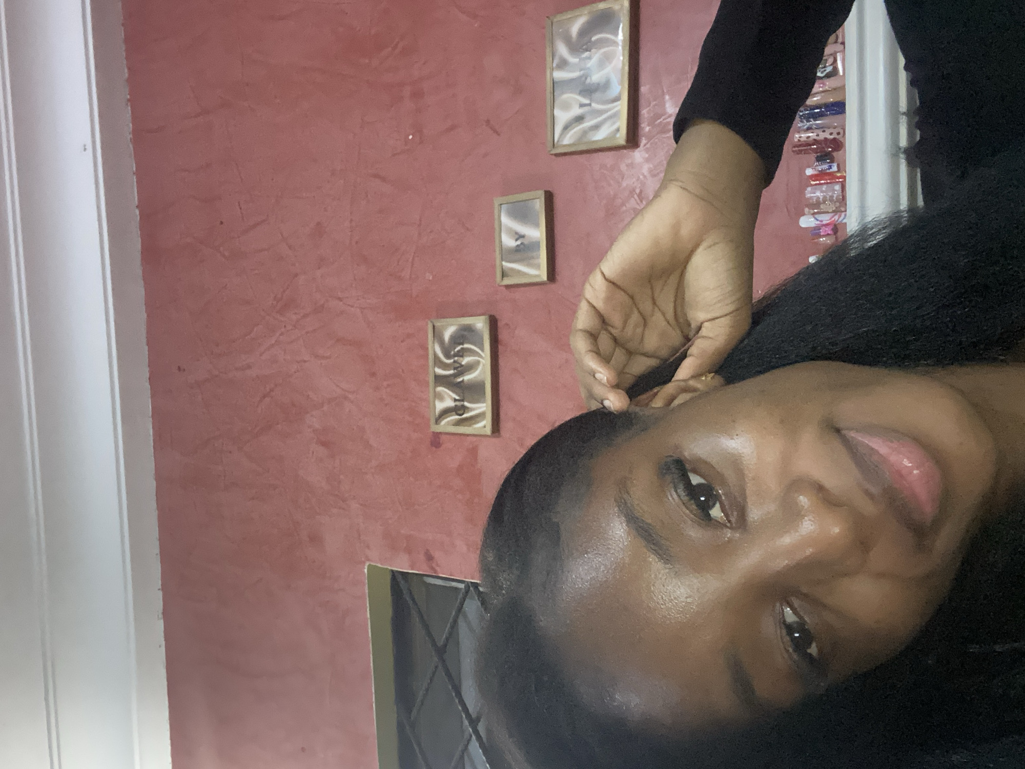 The image shows a close-up of a young Black woman, likely indoors, as indicated by the plastered red wall and framed pictures on it. She is looking towards the camera with a slight smile. Her hand is raised, touching her ear, and her hair is dark and long. On a shelf behind her, a collection of colorful nail polish bottles is visible, some with elaborate designs. The overall lighting suggests it might be evening or a dimly lit indoor space. The room has a textured red wall, a white ceiling trim, and a window with a criss-cross grill. There are three small framed pictures with abstract art or text on the wall. The image does not provide any specific visual cues that can be used to determine the location within the city of Jos, Nigeria.