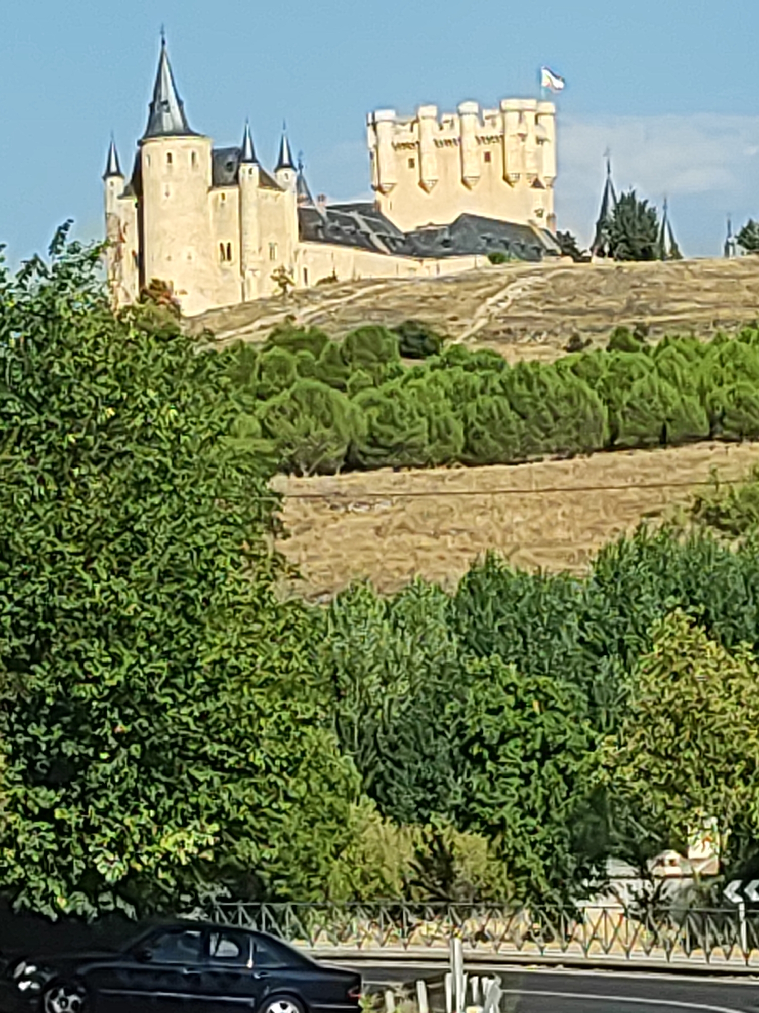 This image captures a panoramic view of the Alcázar of Segovia, Spain, a historic castle situated on a rocky promontory. The castle, constructed primarily from stone, features distinct conical turrets and battlements, with a flag flying from one of its towers. The structure is surrounded by lush green trees and vegetation, with a dry, grassy hillside leading up to the castle. In the foreground, a dark-colored sedan is parked on a road, with a decorative fence bordering the roadway. The sky above is clear and blue.