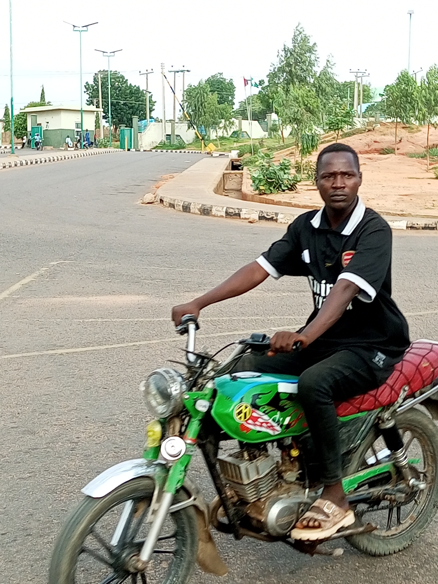 A man is riding a green motorcycle on a paved road in Gombe, Nigeria. The man is wearing a black t-shirt with white trim and black pants. He has on brown sandals. The motorcycle has a red seat and appears to be decorated with some sort of design on the fuel tank. In the background, there are trees, streetlights, and a building with a green gate. There are also several other people and motorcycles visible in the distance, suggesting a public area. The road has white lane markings and a concrete curb. The sky is overcast.