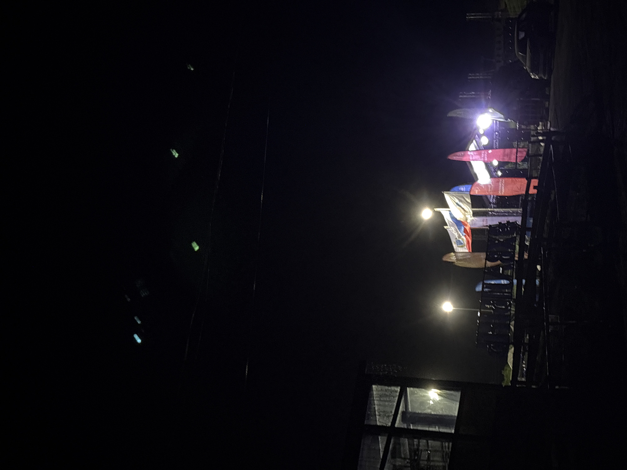 At night in Khvalynsk, Russia, a row of flags is visible under streetlights. A building with a window is partially visible in the foreground. In the distance, a vehicle is parked. The setting is dark with minimal lighting.