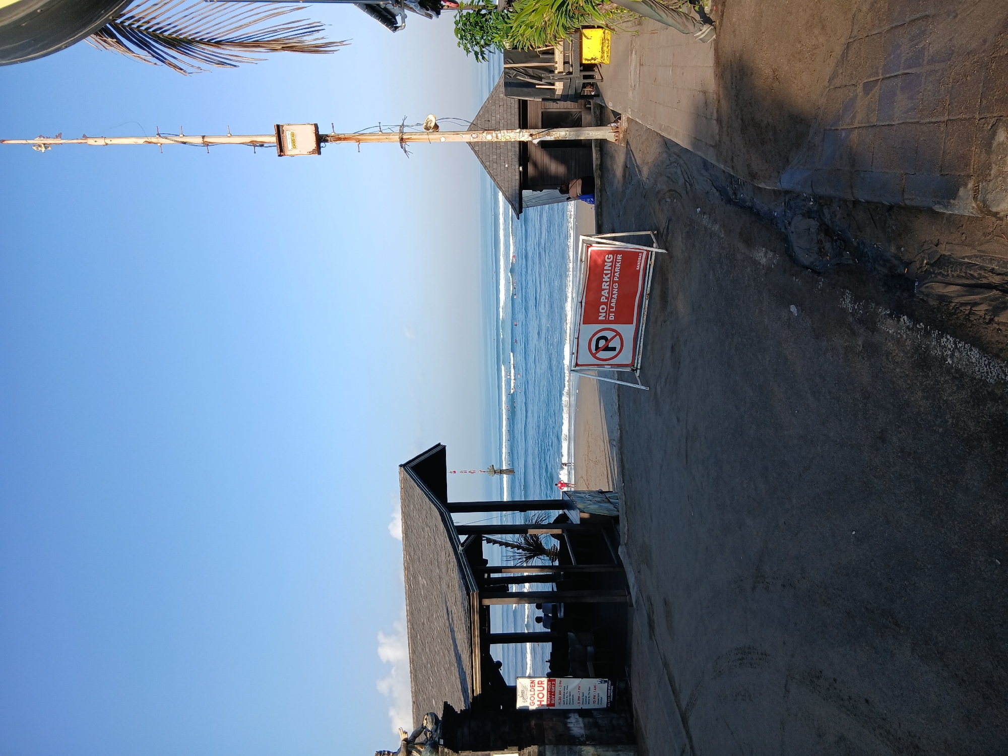 The image depicts a beachside scene in North Kuta, Indonesia, likely during the early morning given the clear blue sky and calm ocean. In the foreground, a sandy area transitions to a paved walkway on the right. A prominent "NO PARKING DI LARANG PARKIR" sign stands on the left, with a smaller "SANDBAR" label below it. Further left, a thatched-roof structure, possibly a beachside cafe or bar, is visible. Several people are seated inside this structure, and two individuals are walking on the beach in the background. The ocean displays gentle waves, with numerous surfers scattered across its surface. To the right of the beach, a tall, slender antenna tower with various attachments and a small box at its base is a notable feature of the landscape, alongside some tropical foliage. The overall impression is of a tranquil, tourist-oriented location.