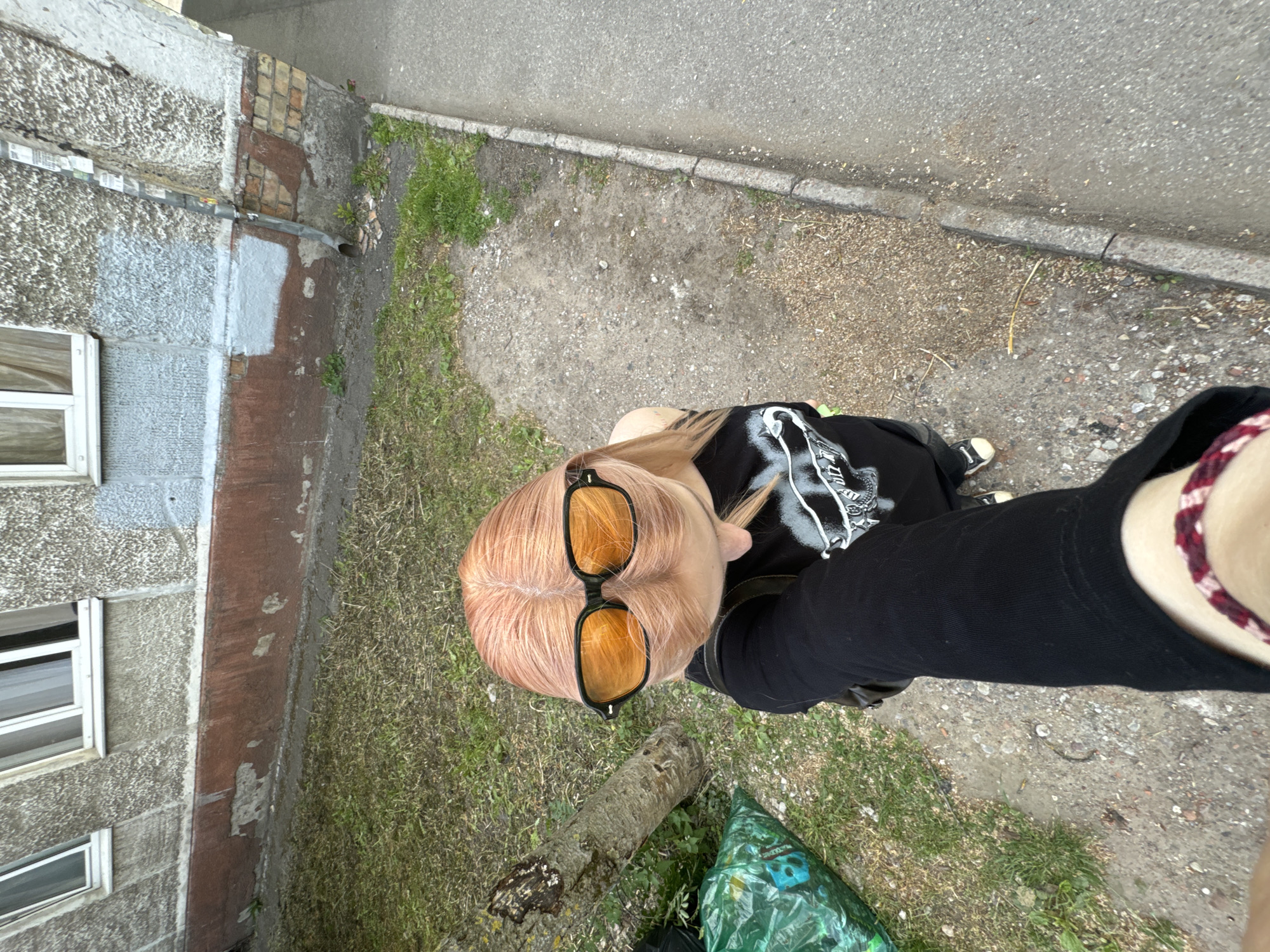 The image is an overhead selfie of a person with light orange hair wearing black-framed sunglasses with orange lenses. They are wearing a black long-sleeved shirt with a white graphic design on the front. The person's left arm is extended towards the camera, displaying a bracelet with maroon and white woven threads.
The setting appears to be outdoors in a somewhat urban environment. A multi-story building with rectangular windows is visible on the left side of the frame. The ground is composed of a mix of concrete, dirt, and grass. A small log lies on the ground with a green plastic bag nearby. A patch of weeds grows between the sidewalk and the building. The building appears weathered and shows signs of repair work in the form of patched sections.
