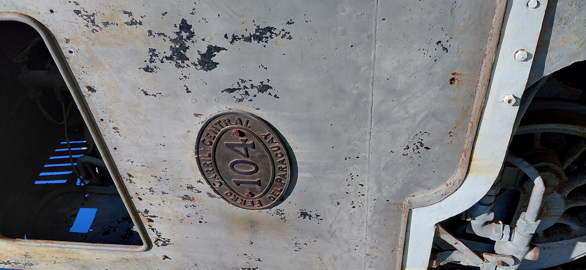 The image shows a close-up of a rusted, grey train car with an oval plaque on its side. The plaque is dark metal and reads "FERRO CARRIL CENTRAL DEL PARAGUAY 104". The number 104 is prominently displayed in the center. The train car shows signs of age and neglect, with peeling paint and rust. The background is blurred, but suggests a sunny day with a bright blue sky visible through some openings in the train car. The context provided by the user states that this image is located in Encarnacion, Paraguay. The scene is static, with no people or active events depicted. The focus is on the train car's exterior and its identifying plaque.