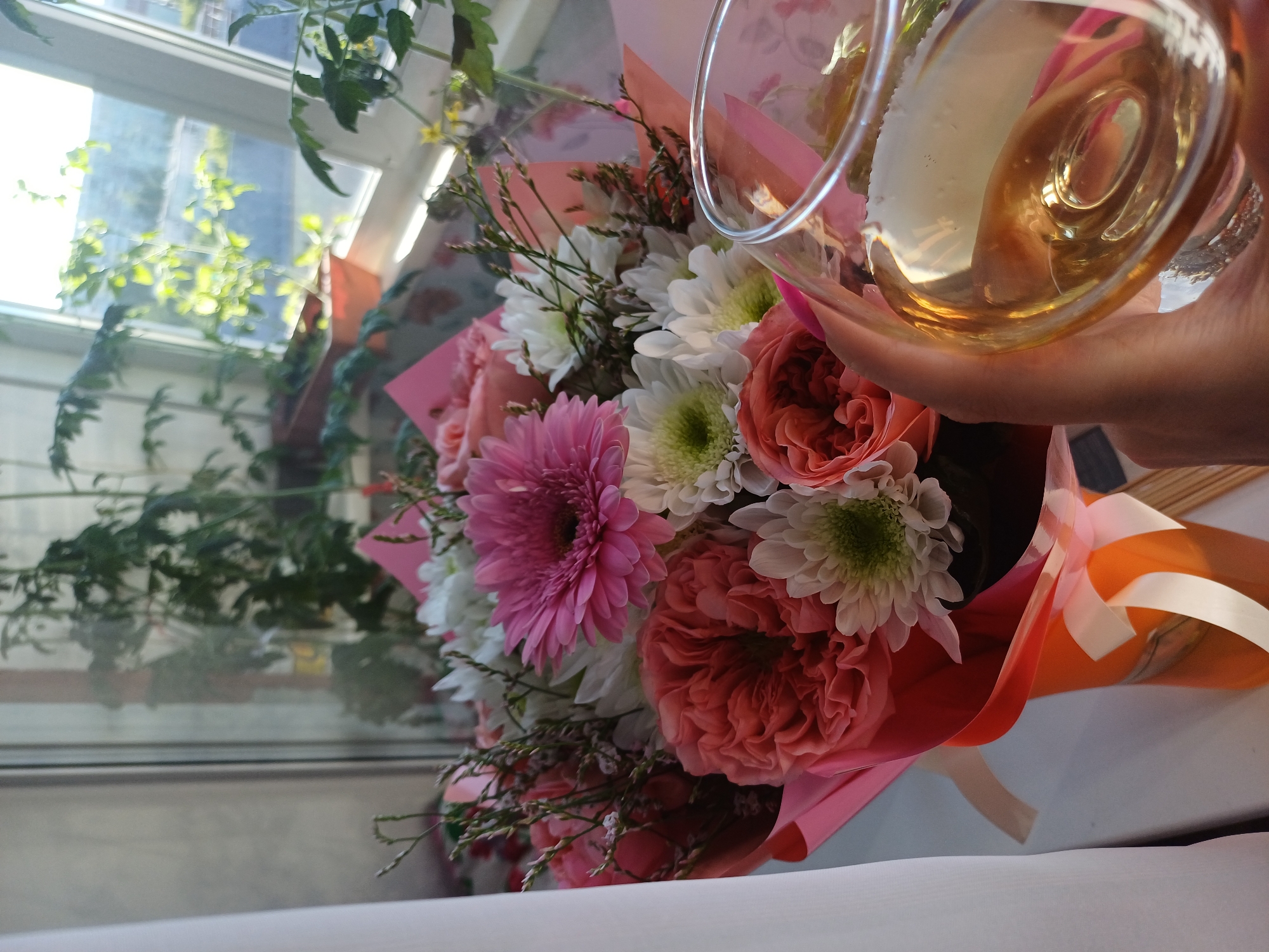 The image shows a close-up of a bouquet of flowers, with a hand holding a glass of light-colored liquid, likely wine. The bouquet contains a variety of flowers in shades of pink, white, and coral. Notable flowers include a large pink gerbera daisy, several fluffy white chrysanthemums, and some coral-colored roses. The flowers are arranged in a vibrant display and wrapped in pink and orange paper.
The background suggests an indoor setting near a window. Green plants are visible outside the window, with sunlight streaming in, indicating it's daytime. The light quality suggests it might be morning or late afternoon. The overall mood is celebratory and serene, with the champagne-like drink and the beautiful floral arrangement. There are no visible text elements in the image.