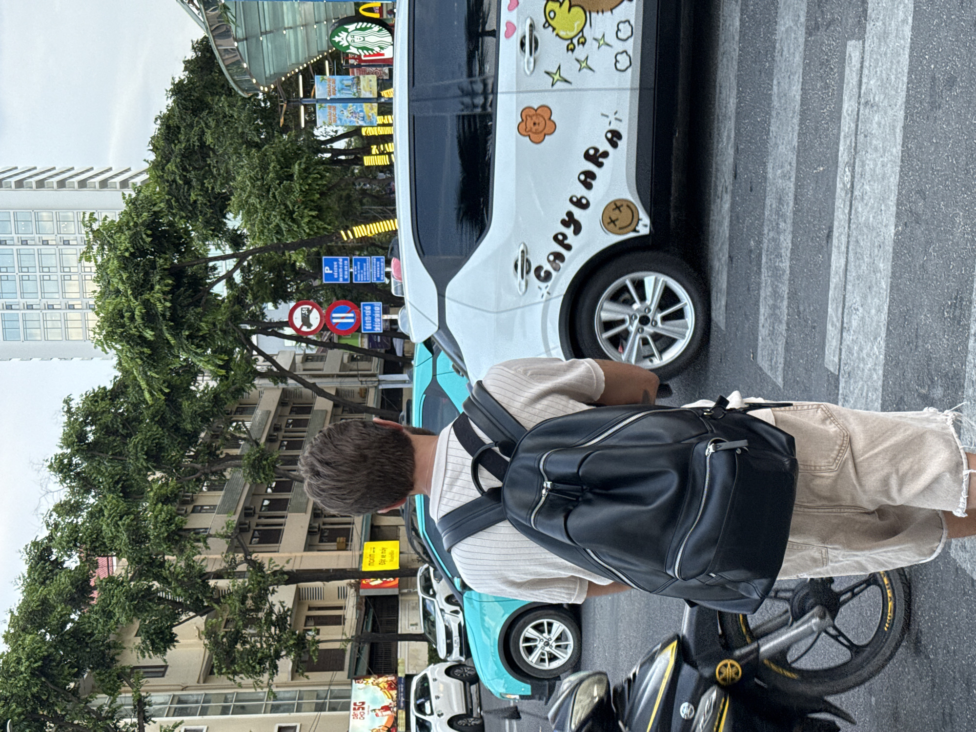 The image depicts a street scene, likely in Nha Trang Ward, Vietnam, as suggested by the context. A young man, seen from behind, stands on a pedestrian crossing, wearing a light-colored short-sleeved shirt and light-colored shorts, and carrying a black backpack. He appears to be waiting to cross the street.
Several vehicles are visible. A teal-colored car is positioned to his left, and a white car, decorated with cartoonish drawings and the text "CAPYBARA," is to his right. A motorcycle is parked on the far left side of the frame.
The background shows multi-story buildings and lush green trees lining the street. Traffic signs, including what appear to be traffic regulations, are present on the left side of the image. The sky is overcast. The overall impression is that of a typical urban street in Southeast Asia.
