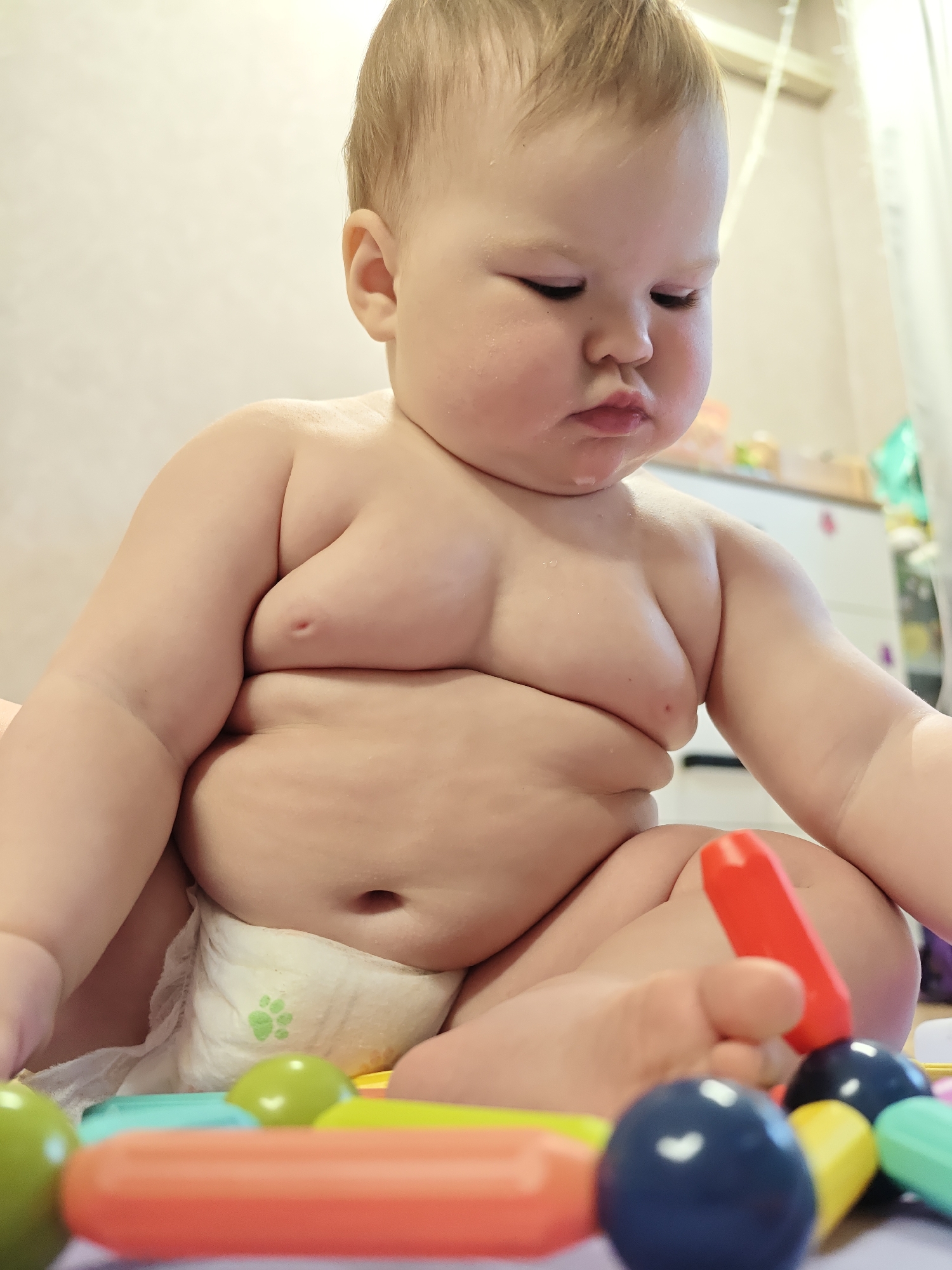 A close-up shot shows a baby in a diaper playing with colorful toys. The baby has light skin, blond hair, and is looking down at the toys. The baby is sitting in a diaper with a green paw print design. The foreground features an array of colorful toys, including spheres and rods in red, blue, yellow, and green. The background shows a wall and part of a white dresser. There is no verifiable evidence to associate the image with the city of Chaikovsky, Russia.