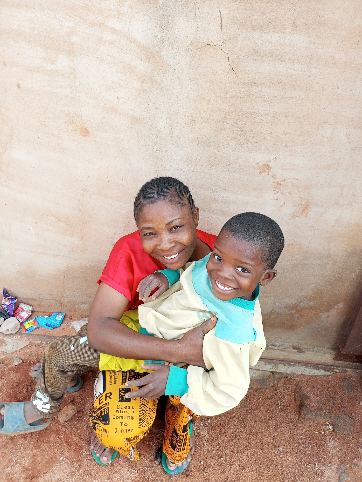 A medium shot captures a woman holding a child in front of a tan-colored, rough-textured wall. The woman, on the left, wears a red t-shirt and brown pants with white markings. She has her hair braided and is smiling at the camera. The child, held by the woman on the right side of the frame, wears a cream-colored long-sleeved shirt with light blue trim. The child is also smiling at the camera. The ground is covered in reddish-brown dirt and small pebbles. Scattered to the left of the woman are several colorful packets of snacks and some small rocks. The wall appears weathered with some cracks and stains. The scene is lit by natural light.
