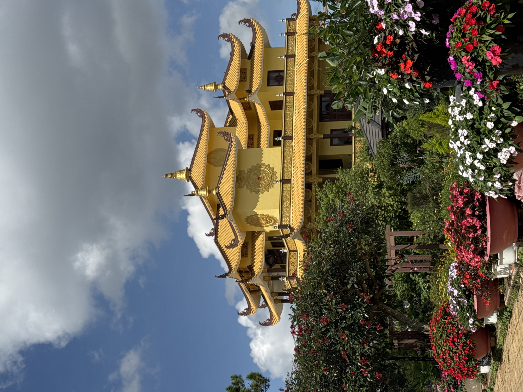 The image depicts a multi-tiered, yellow building with ornate golden roof details, situated in Da Lat, Vietnam. The building is partially obscured by lush vegetation, including trees and potted plants with red, white, and purple flowers. The sky is visible in the background, featuring a mix of blue sky and white clouds. The image is oriented vertically, potentially rotated.
